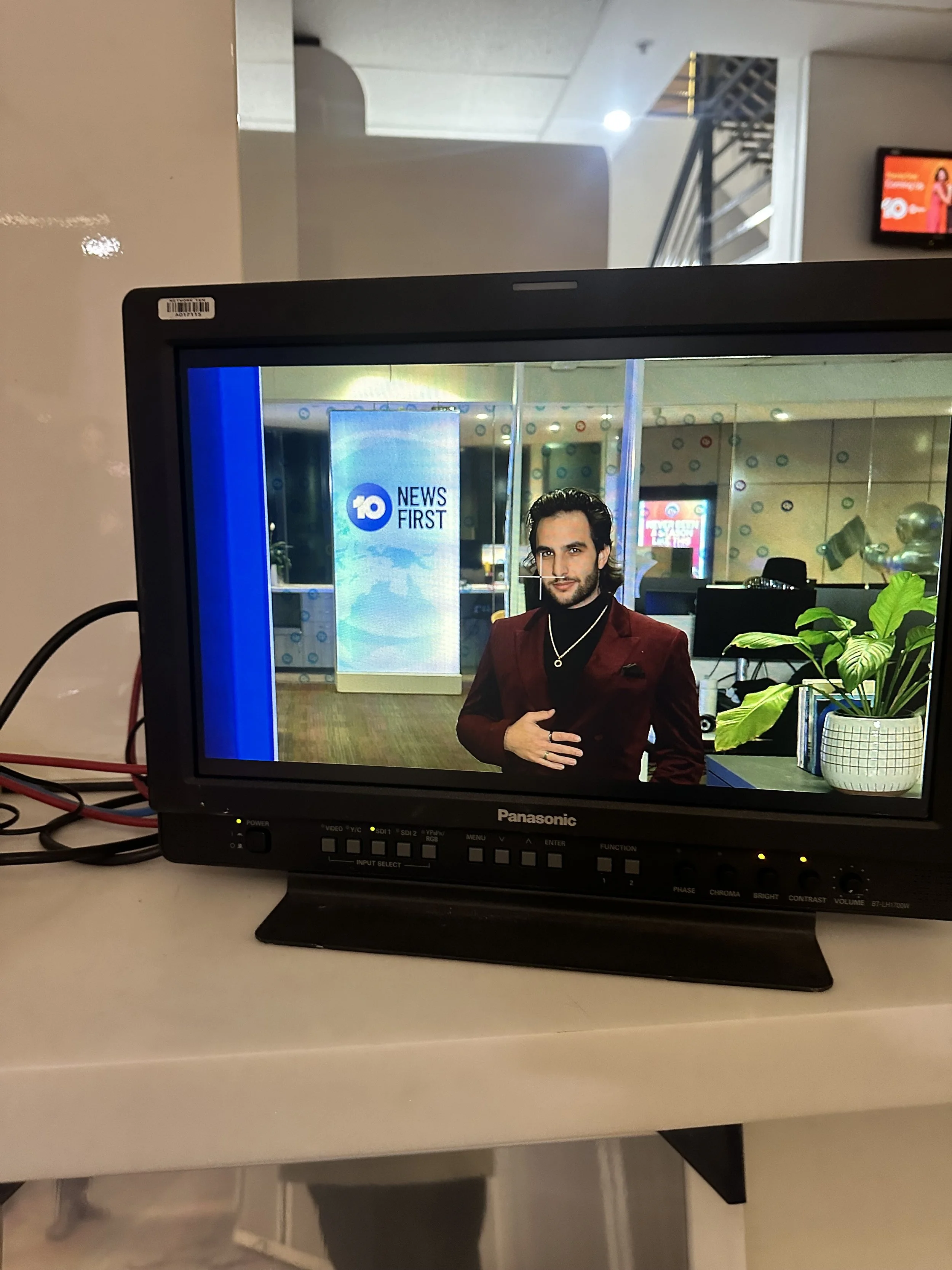 Monitor displaying a news anchor with a logo reading '10 News First' in the background, a plant on the right side, part of a newsroom visible behind, and a staircase with a metal railing in the upper background.