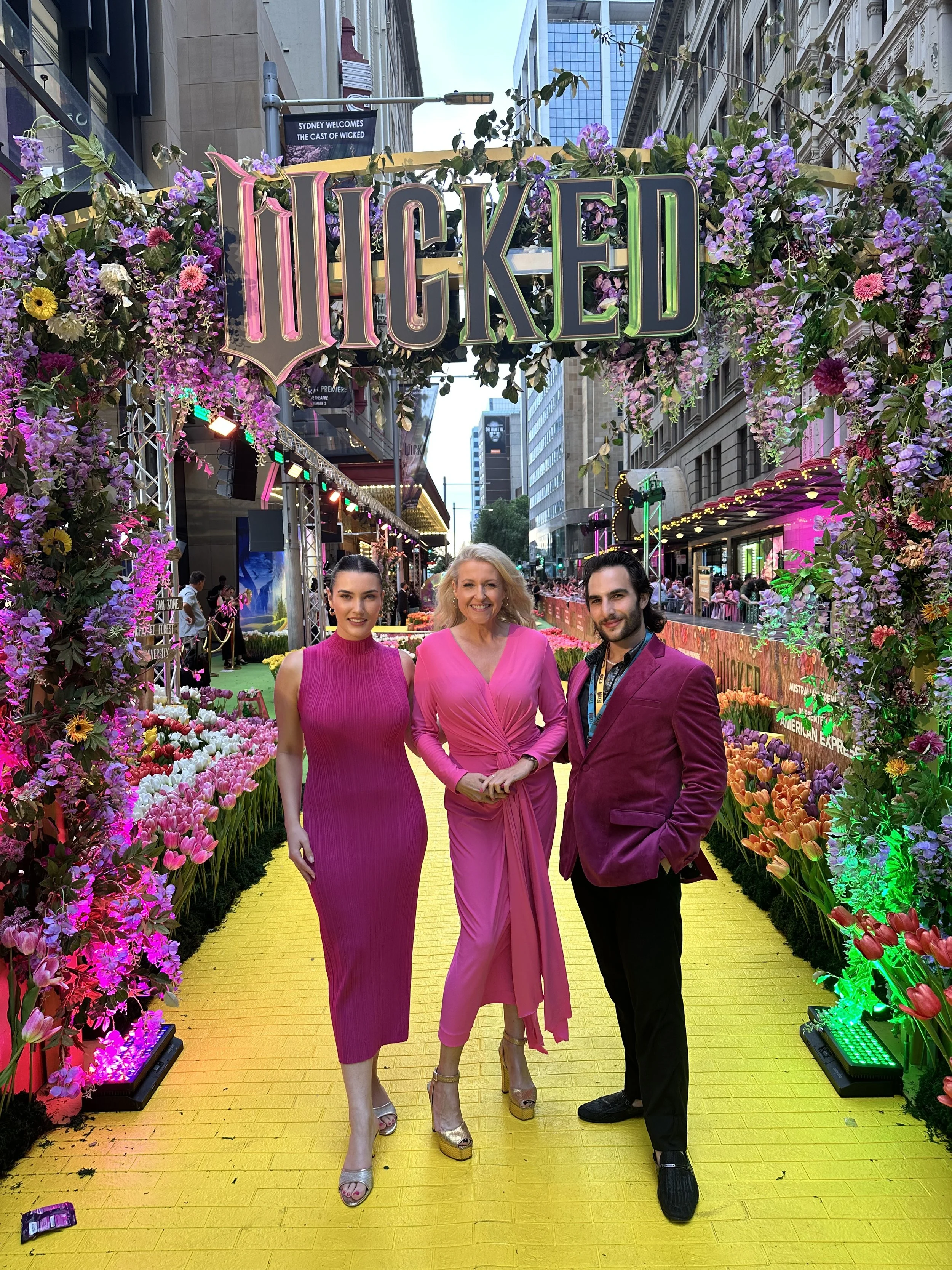 Three people, two women and one man, standing on a bright yellow floor at a floral event in a city street, surrounded by colorful flowers and decorations, with a large sign that says 'HOOKED' overhead.