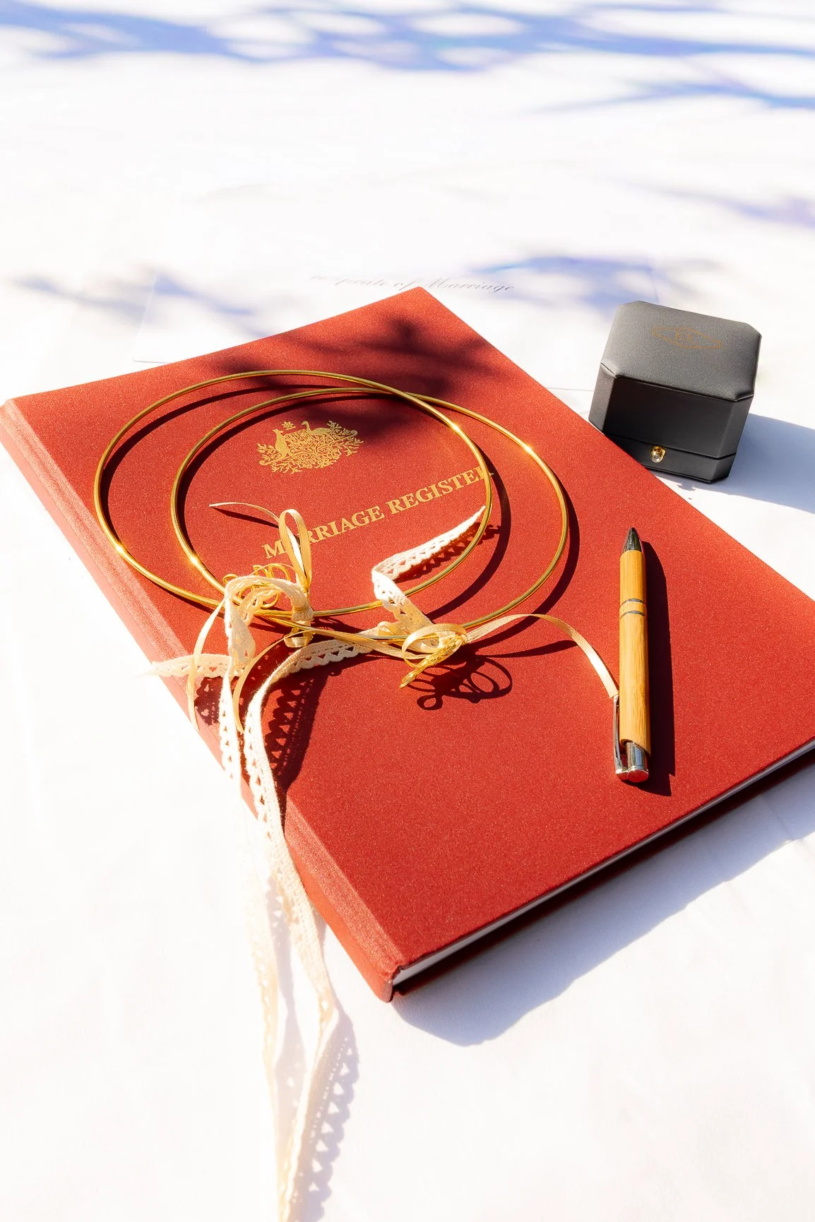 A red marriage register book with a gold crest and text, wedding rings, a gold pen, and a small black box on top of white paper outdoors.