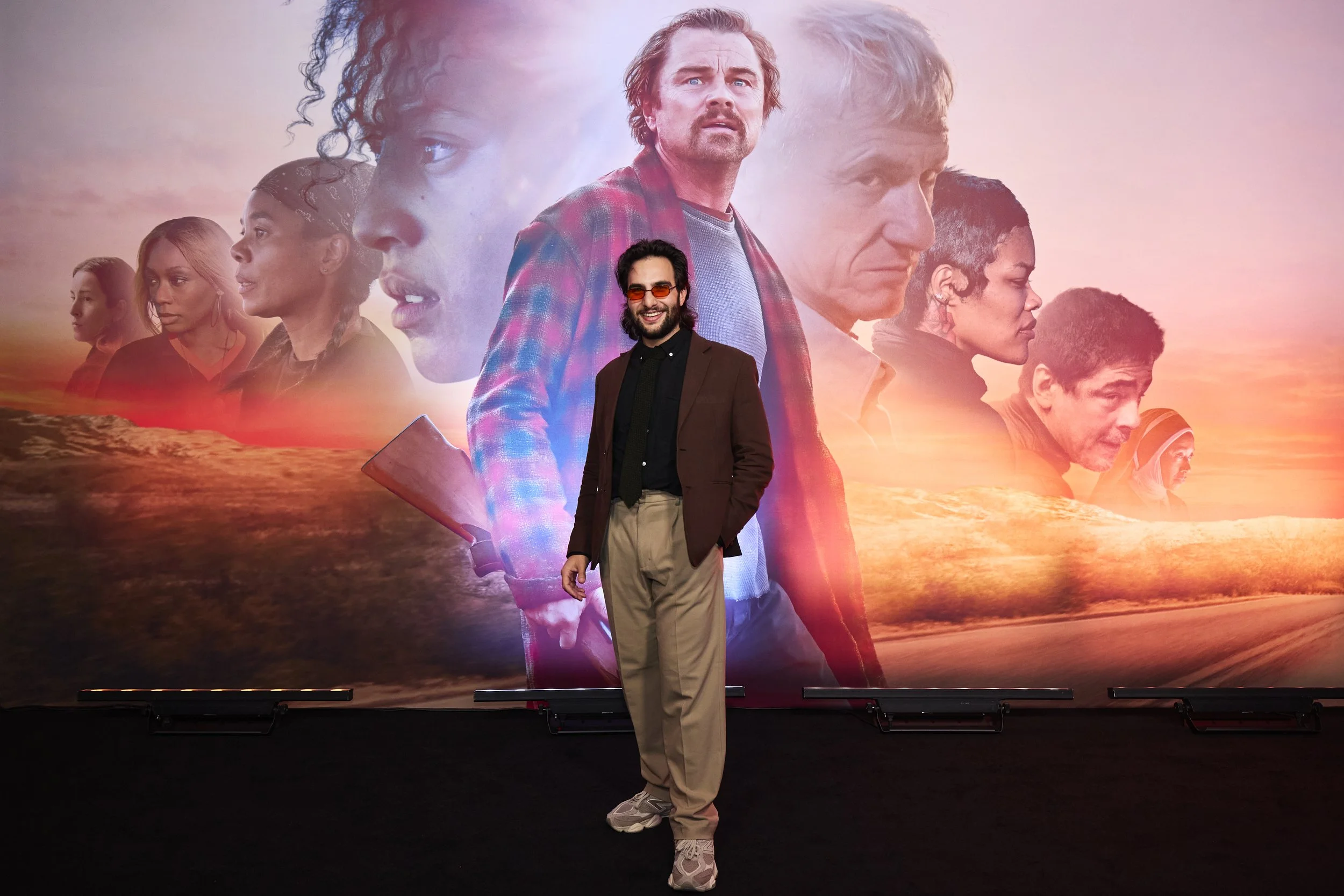 A man with glasses and a beard wearing a brown blazer, black shirt, and beige pants standing in front of a movie poster with multiple faces, vibrant sunset colors, and two comic book-style comic bubbles.