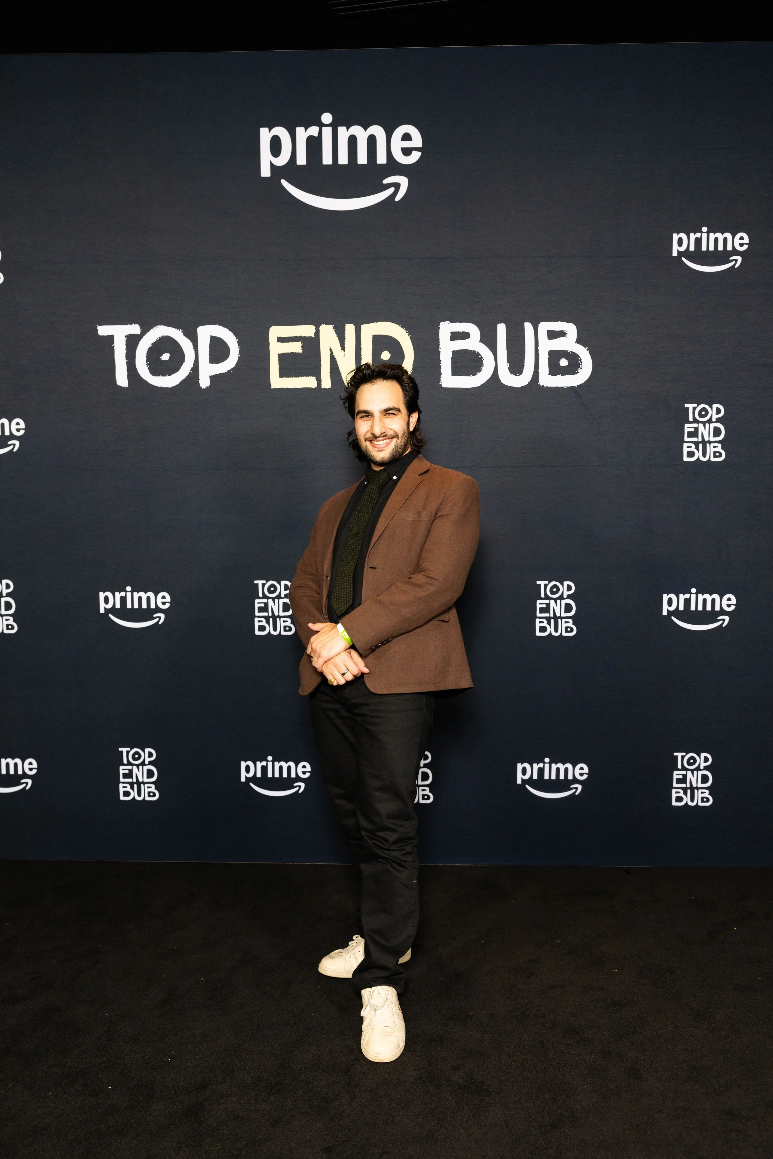 A man standing with arms crossed in front of a dark background with the words 'Top End BUB' and 'prime' logos repeated. The man has dark hair, a beard, and is wearing a brown blazer, black shirt, black pants, and white sneakers.