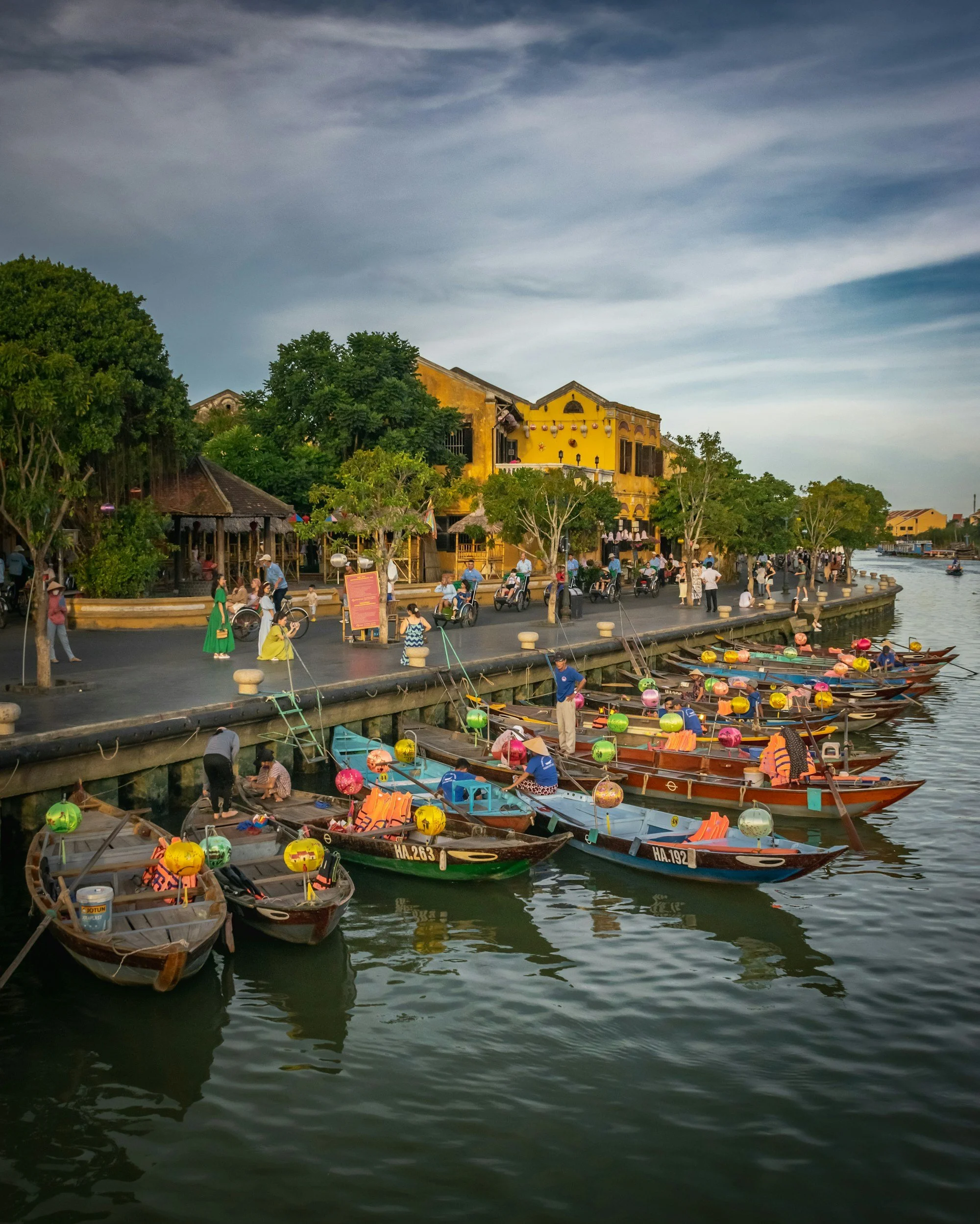 People walking, biking, and sitting along a riverside walkway with colorful boats docked in the water, yellow buildings, and trees, under a partly cloudy sky.