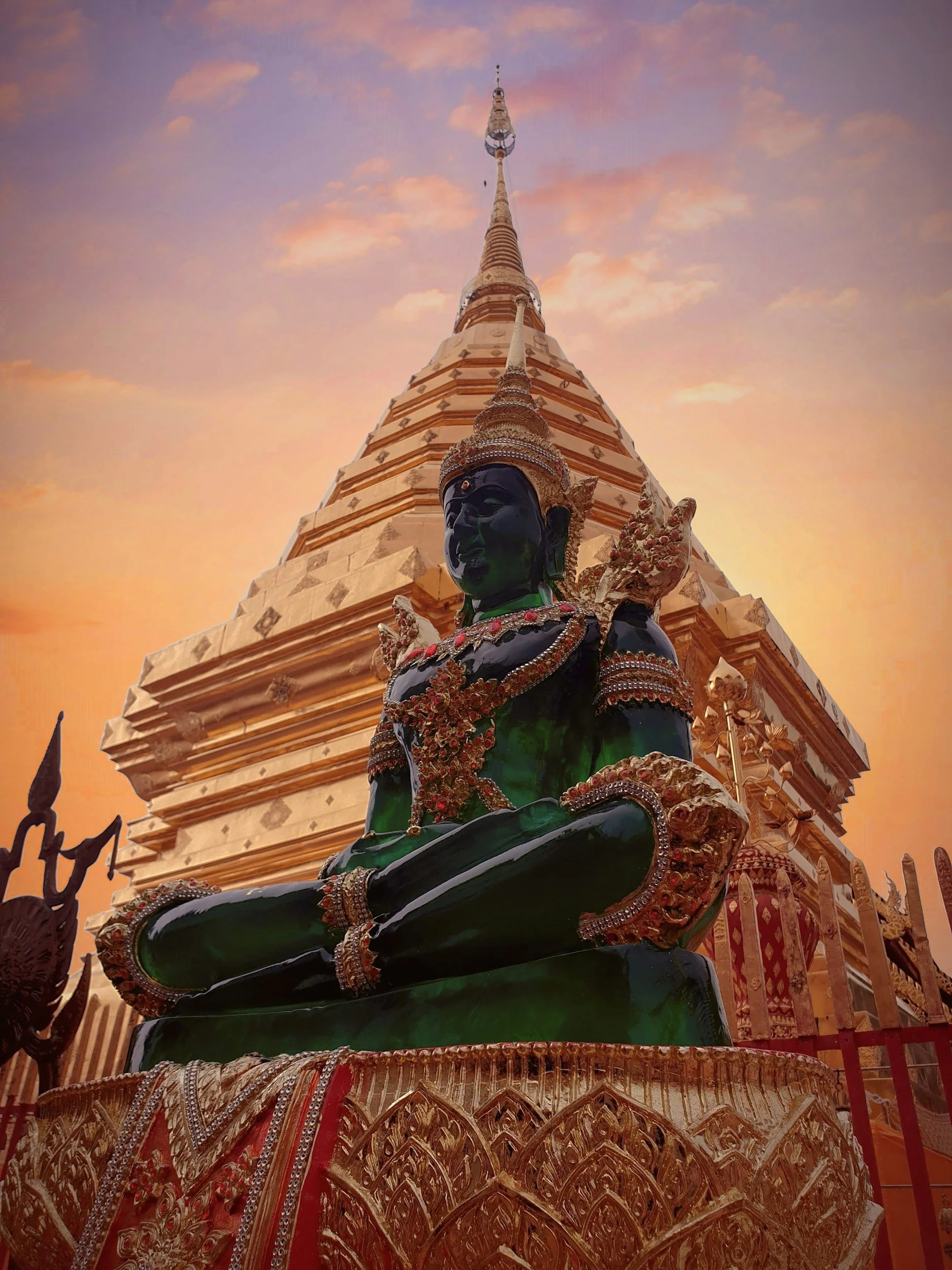 A large ornate temple spire with a dark statue of a mythological figure in the foreground, adorned with jewels and intricate decorations, against a sunset sky with clouds.