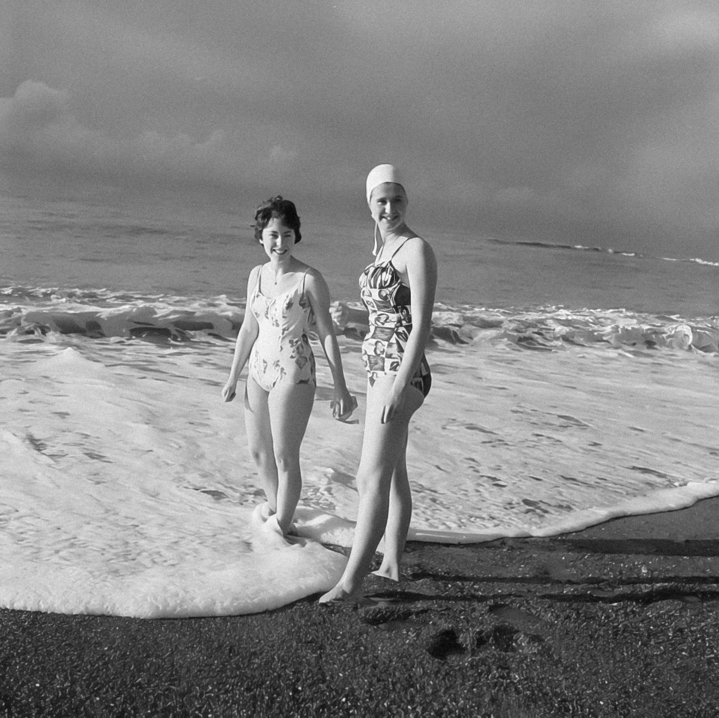 Two women walking on the beach with waves in the background, black and white photo.