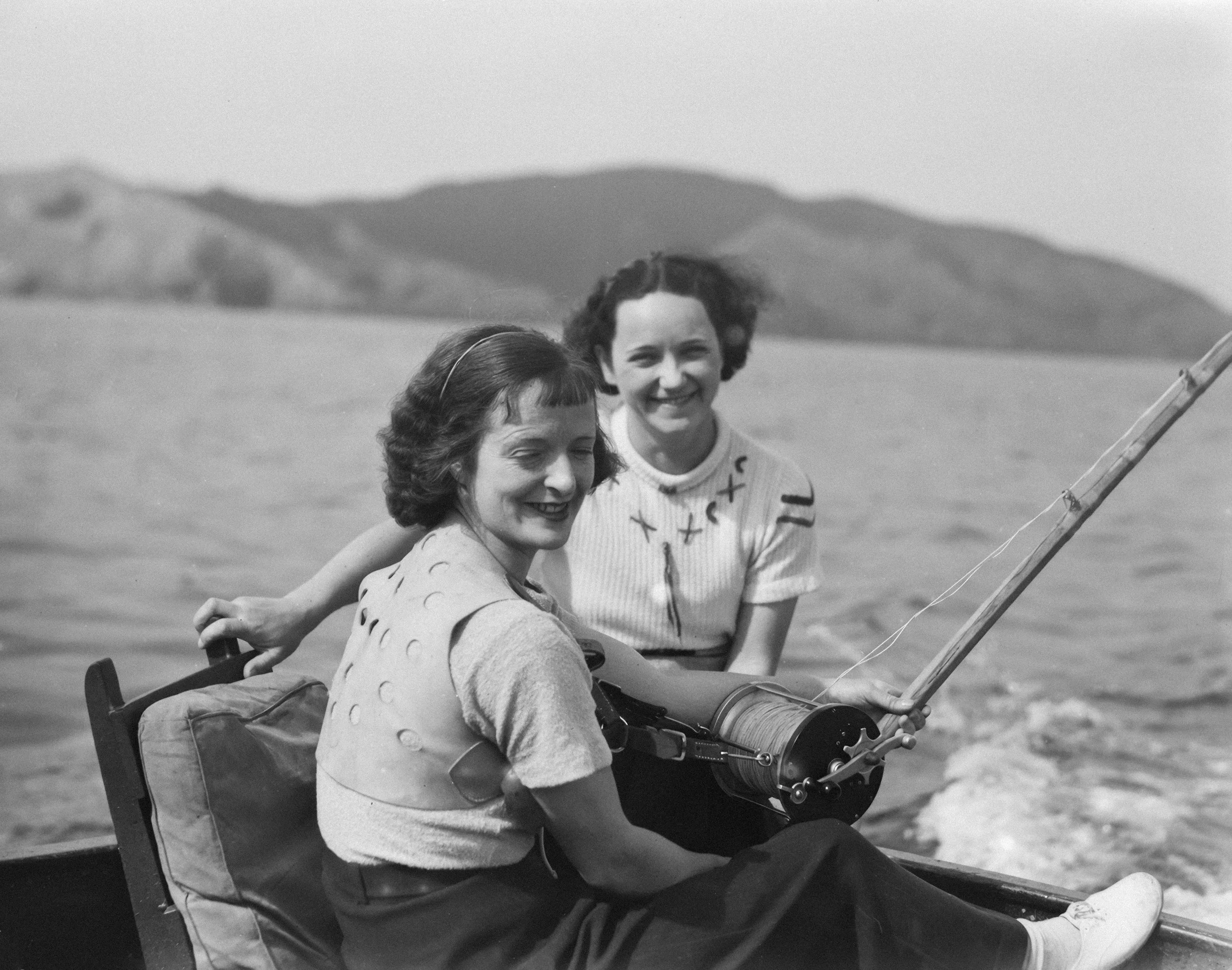 Two women smiling outdoors, with one seated and the other standing, holding a camera mounted on a large tripod, with mountains in the background.