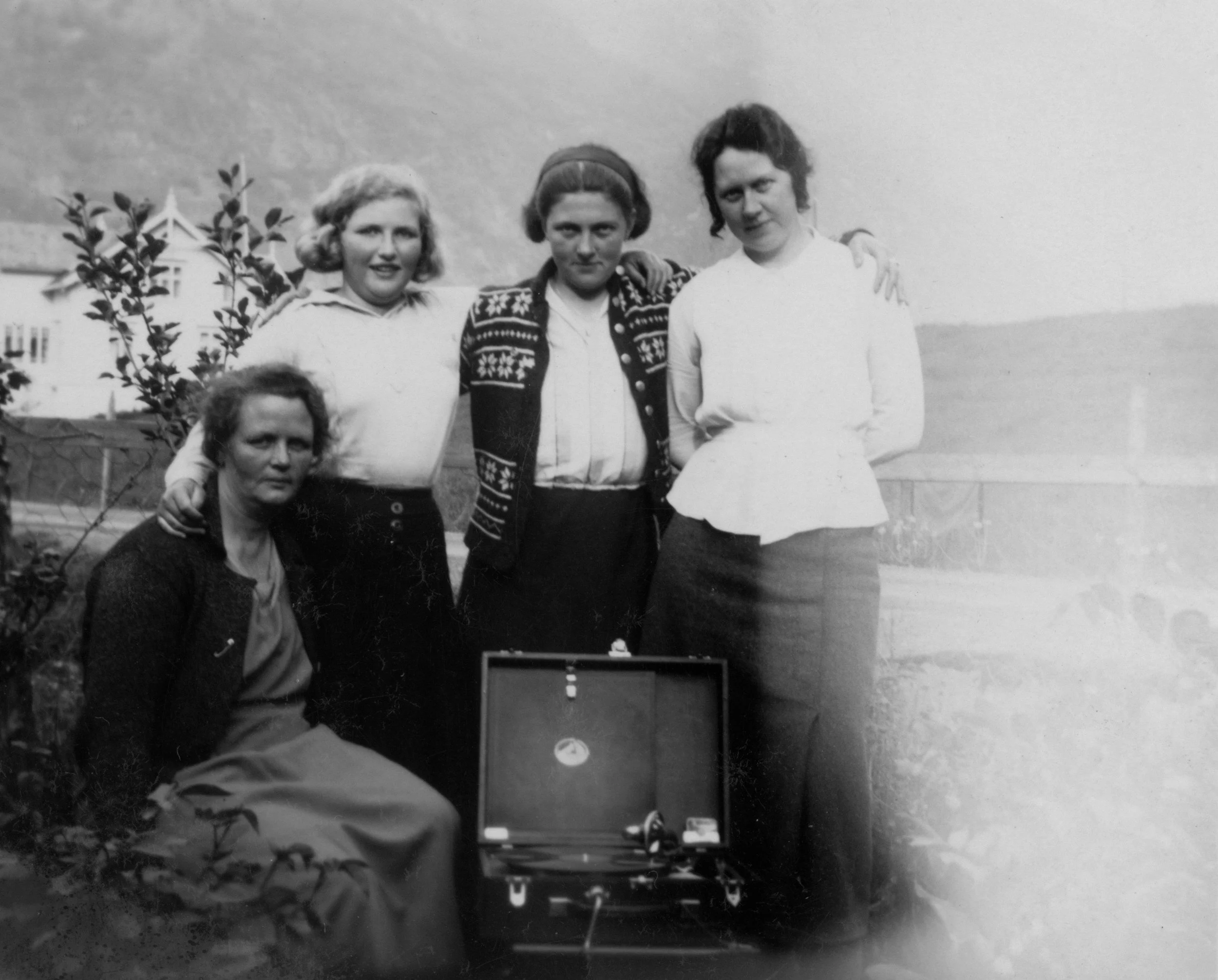 Black and white photo of three women standing outdoors near a body of water, with a building in the background. One woman is squatting in the foreground, and the other two are standing behind her. All three women are dressed in early 20th-century clothing, and the woman in the center is wearing a patterned sweater, while the woman on the right has her arm around the center woman's shoulder.