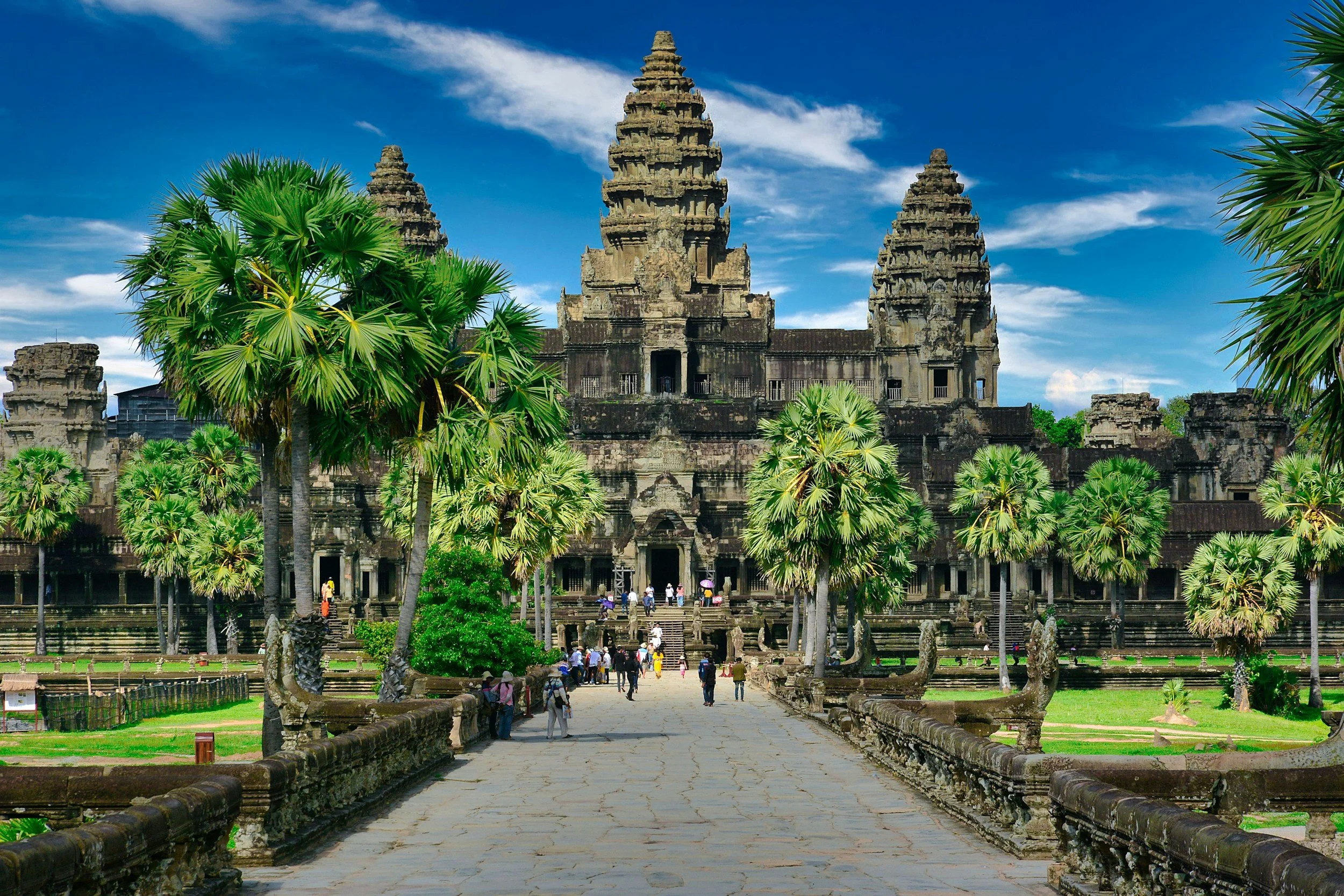 Ancient stone temple with three tall towers, surrounded by lush green palm trees, blue sky with a few clouds, and visitors walking along the pathway leading to the temple entrance.