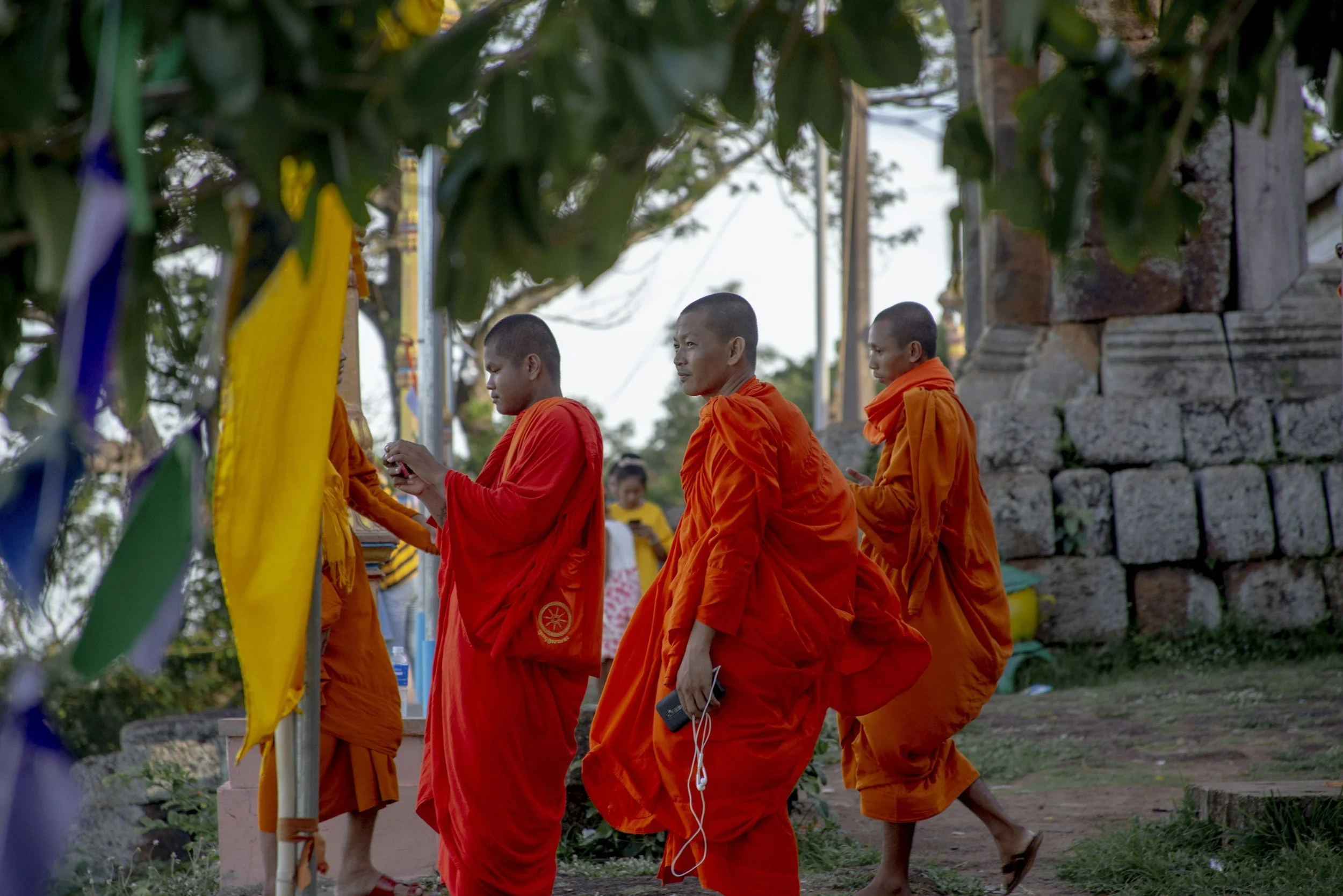 Four Buddhist monks in orange robes walk in a row outdoors, some are praying and some are with their eyes closed, near a stone wall and trees.