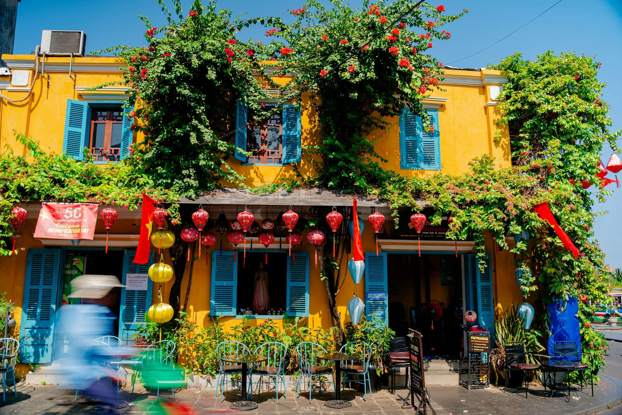 Yellow building with blue shutters, decorated with red lanterns and featuring lush green vines and trees in front under a clear blue sky.