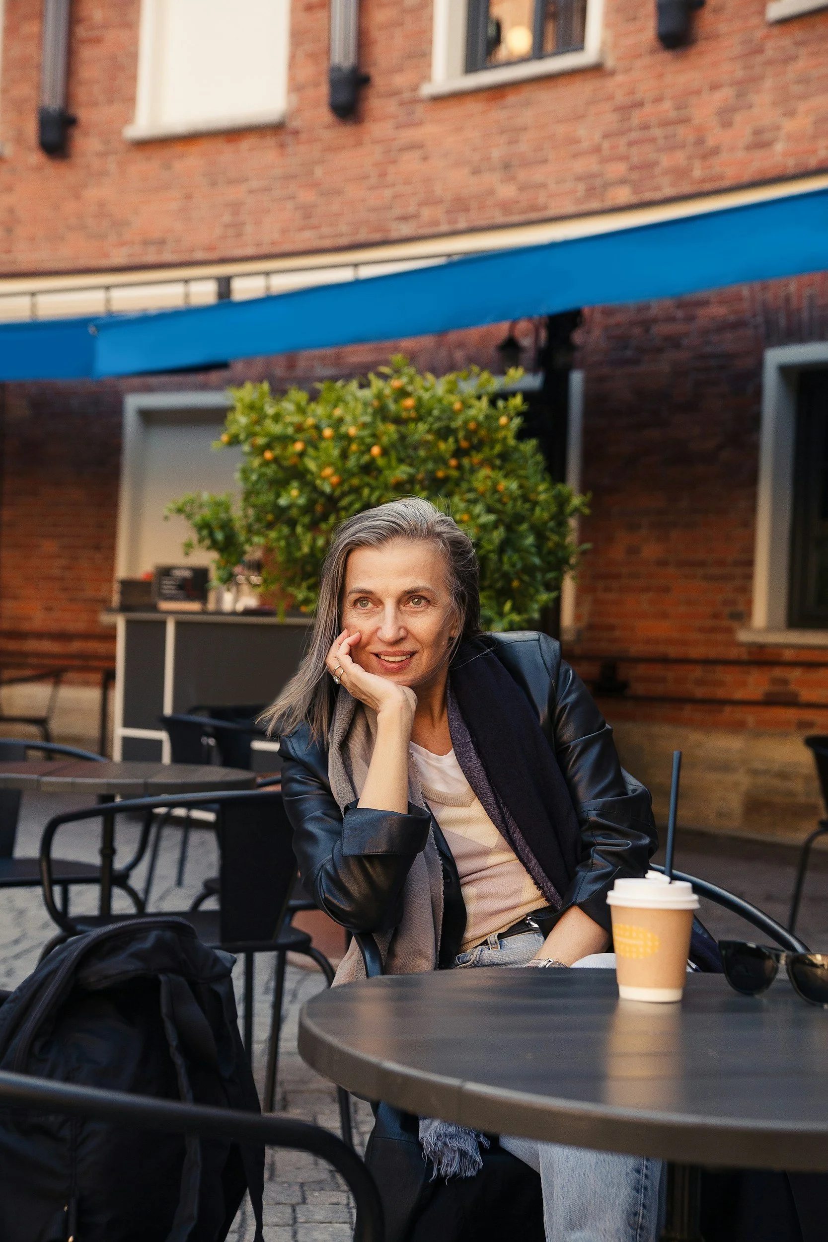 A woman with gray hair sitting at an outdoor café table, smiling and resting her chin on her hand, with a hot coffee in a to-go cup on the table, in front of a brick building and a tree with green and orange leaves.
