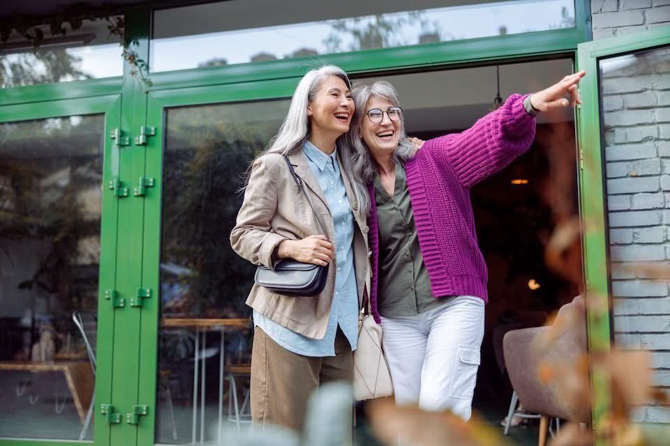 Two women are standing by a large glass door, smiling and laughing. One woman, wearing a beige jacket and blue shirt, has gray hair, and the other woman, wearing a purple cardigan and glasses, is pointing outside.