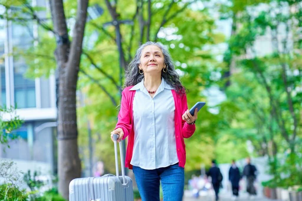 An elderly woman walking outside with a rolling suitcase and holding a smartphone.