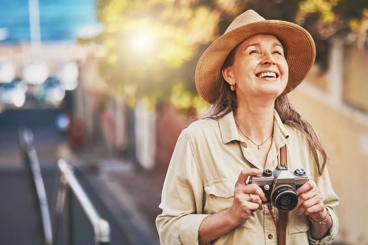 A smiling elderly woman wearing a wide-brimmed hat and light-colored shirt, holding a vintage camera, standing outdoors with blurred trees and sunlight in the background.