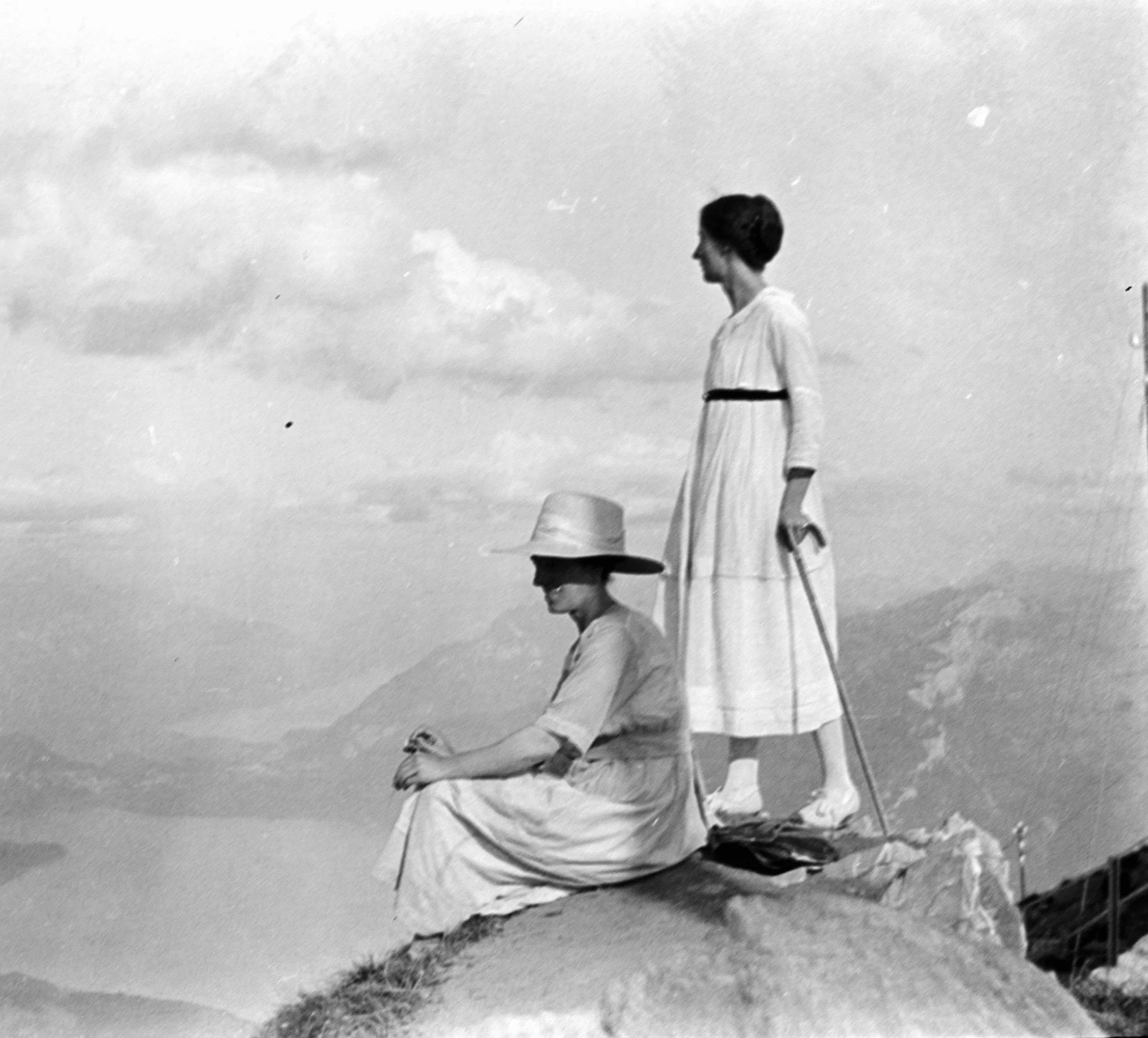 Black and white photo of two women in vintage dresses and hats sitting on a log by water, facing opposite directions, with clouds in the sky.