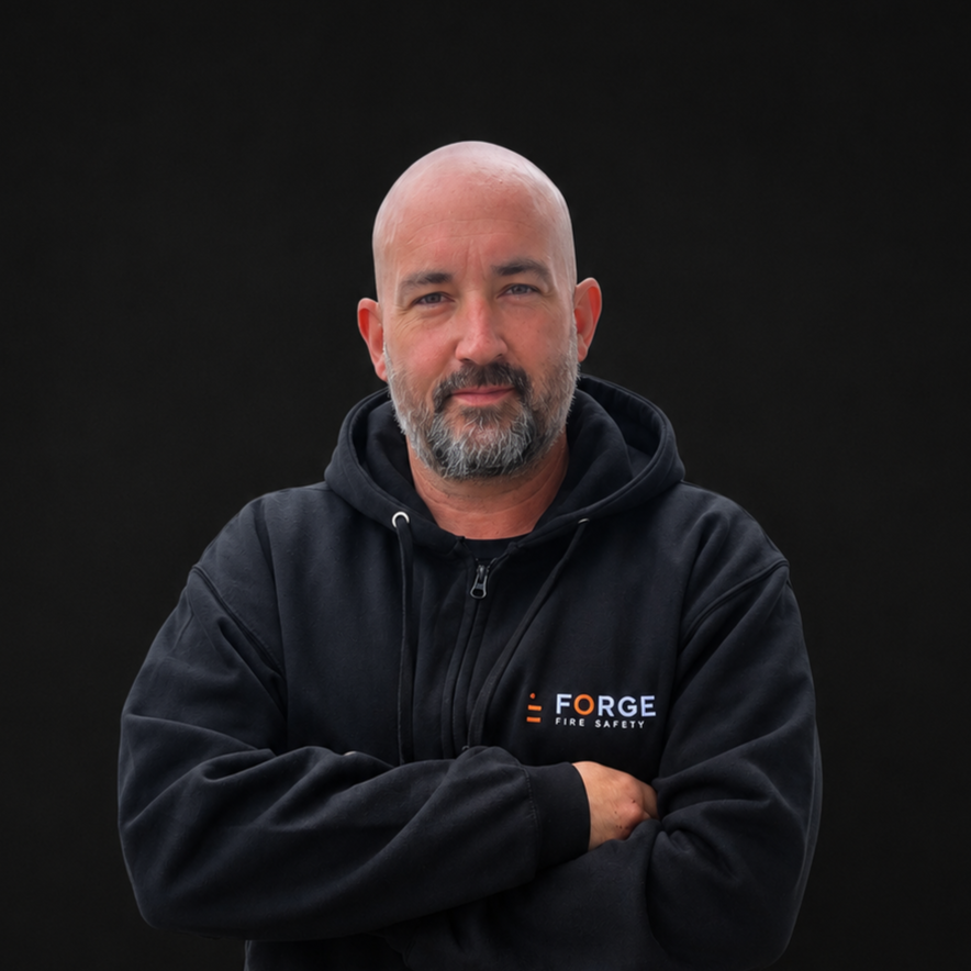 A man with a beard and bald head wearing a black hoodie with 'FORGE FIRE SAFETY' logo, standing with his arms crossed against a black background.