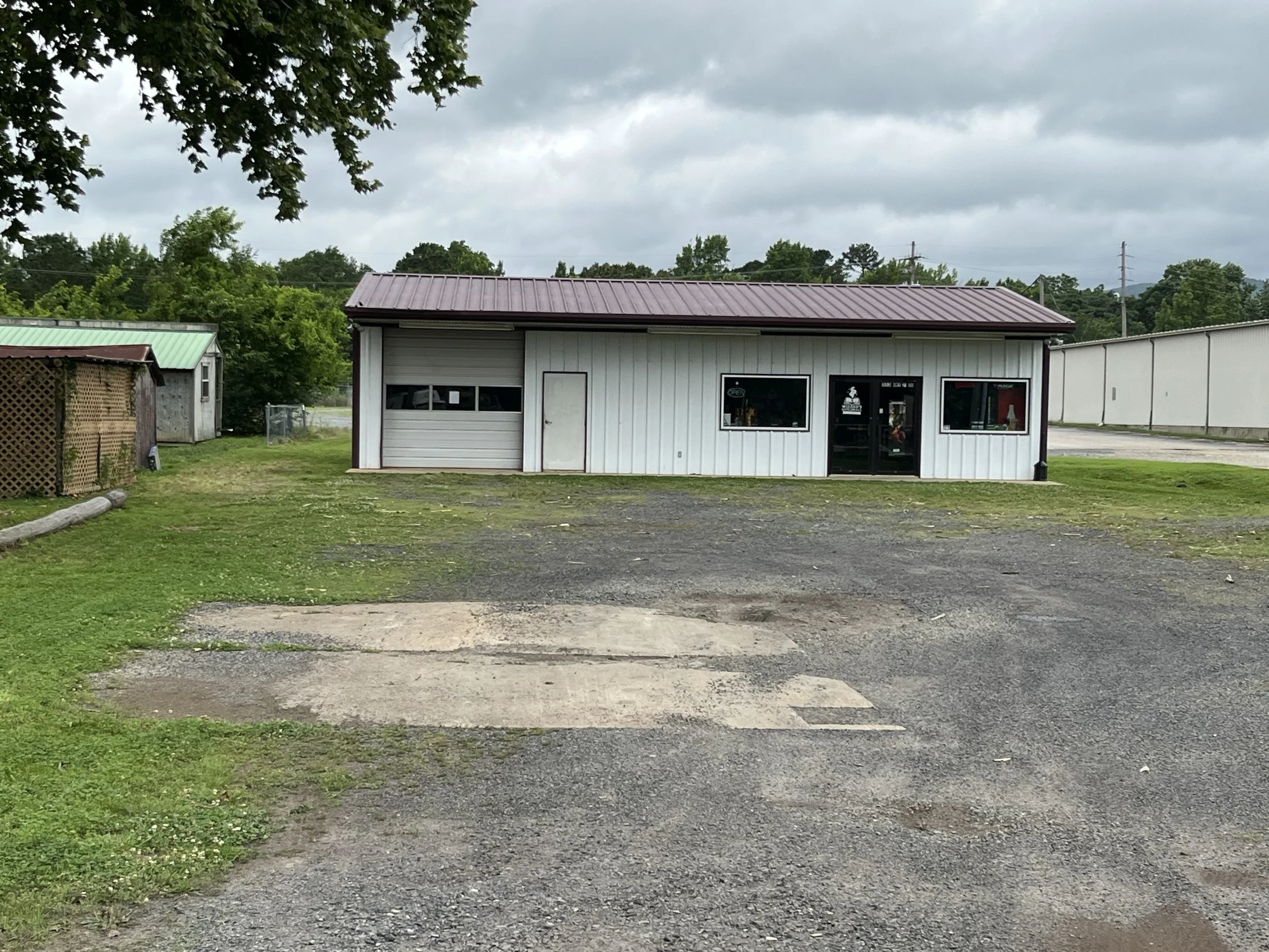 A small white building with a red metal roof, large windows, and a glass door, situated in a semi-rural area with green trees and cloudy skies in the background.