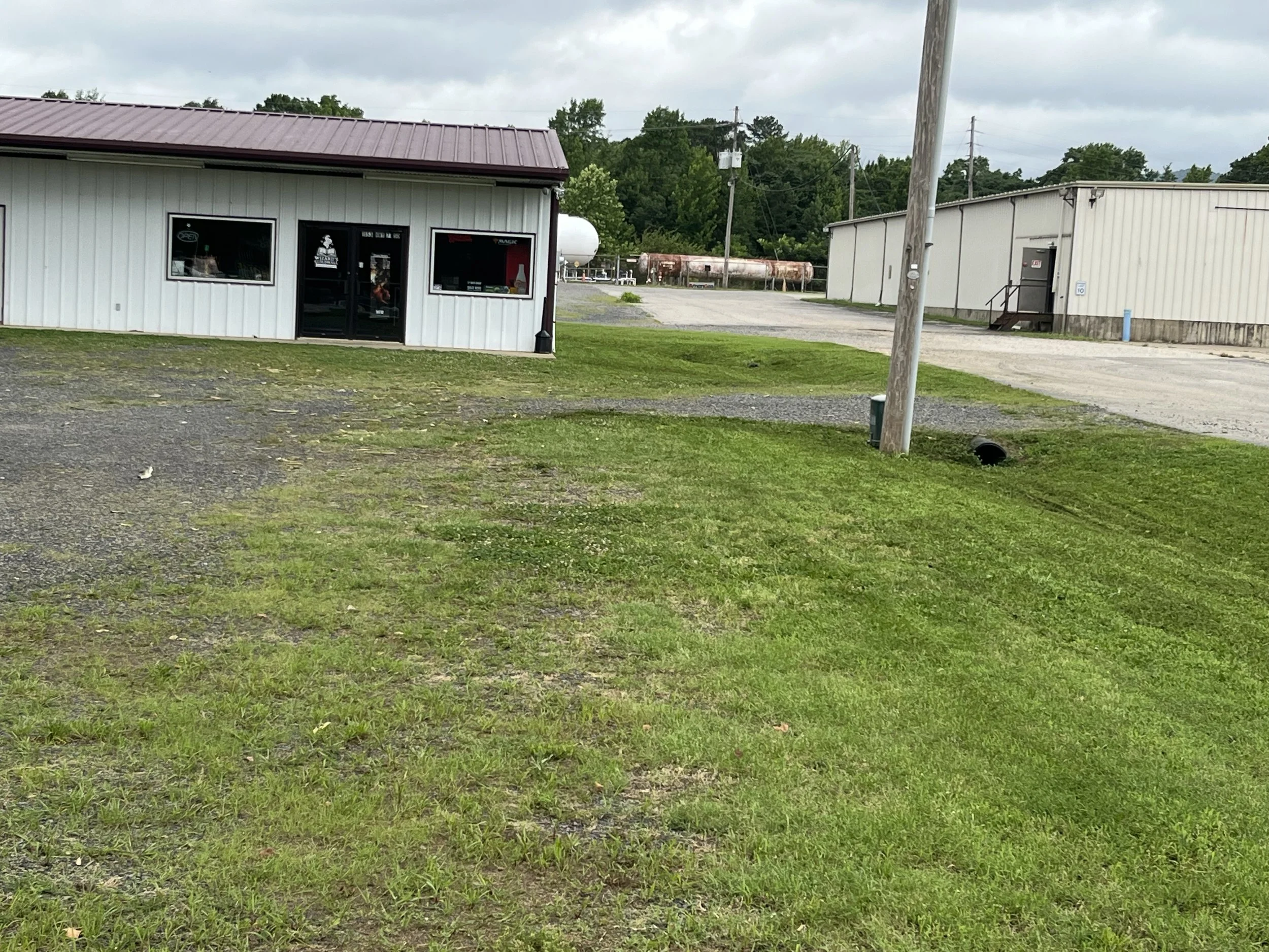 A small commercial building with white siding and a maroon metal roof. There are two windows, one with an open sign and another with various stickers. The door has a small window with the business logo and a sign. To the right, there is a larger industrial building with a side door and steps. Behind, there are trees, utility poles, and a large rusted tank or pipe horizontally laid.
