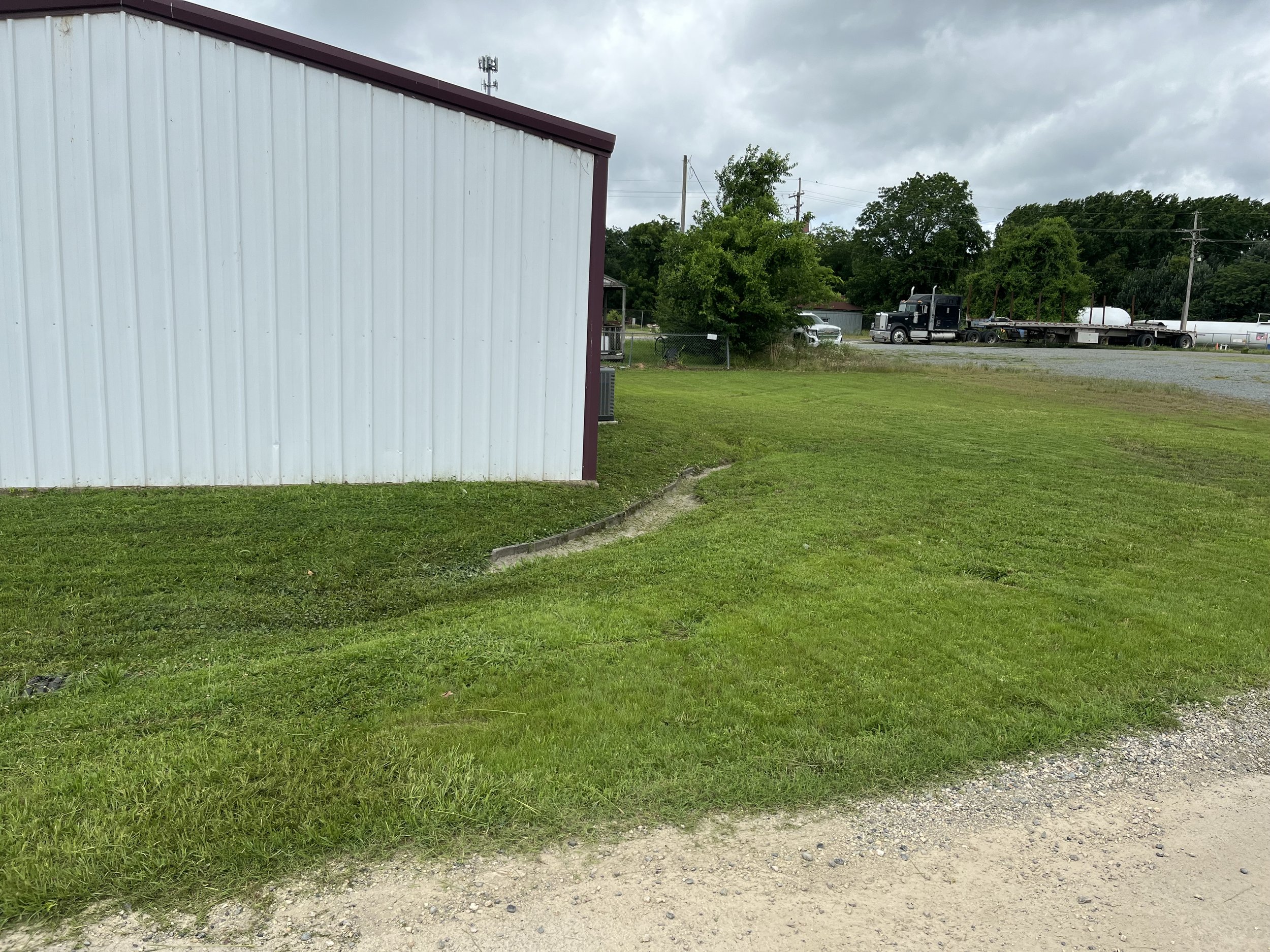 Green grass and dirt path in the foreground, white building with maroon trim on the left, trees and utility poles in the background, dark cloudy sky overhead.