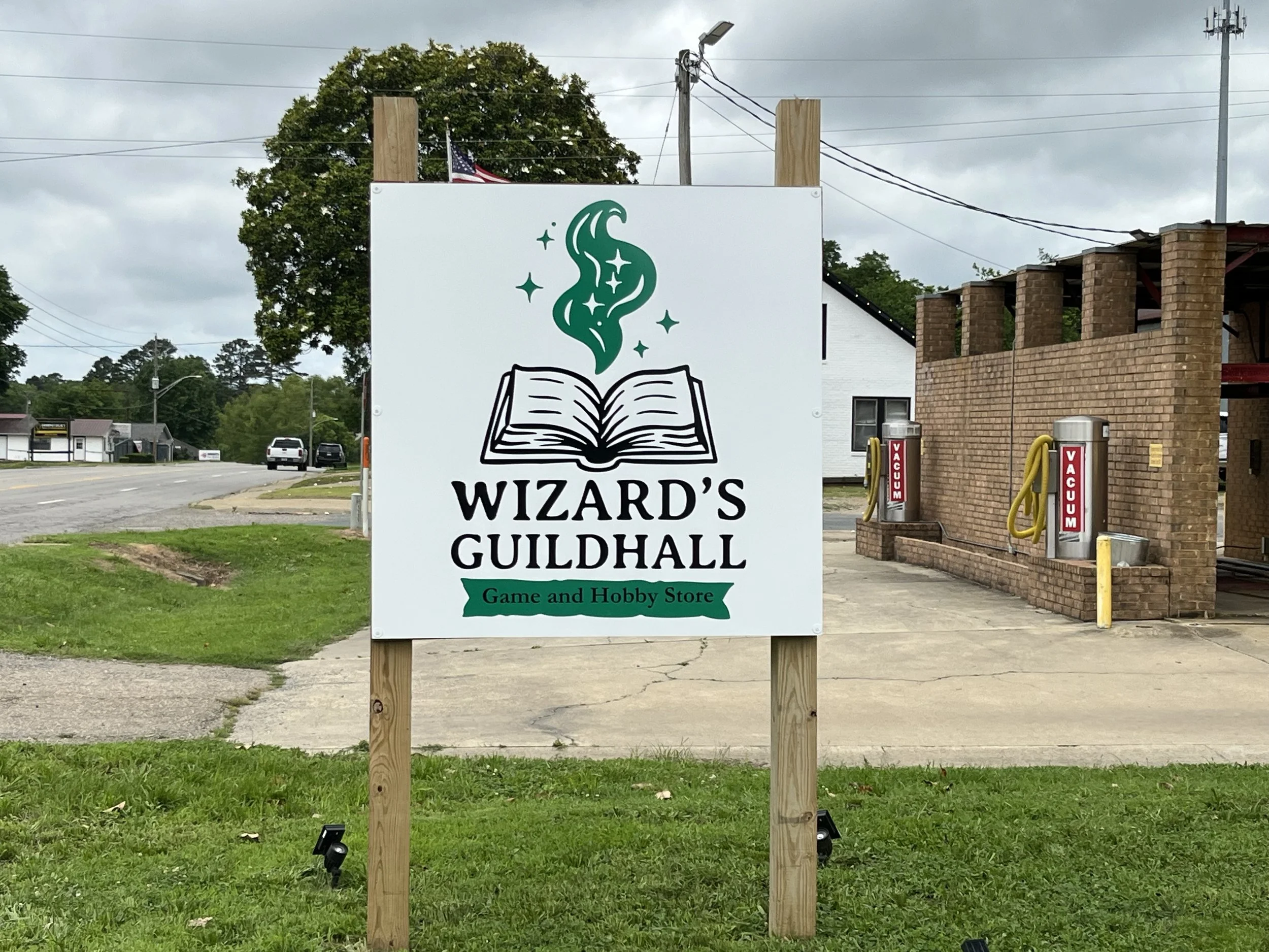 Sign for Wizard's Guildhall, a game and hobby store, with an illustration of a magic book emitting green sparkles and smoke.