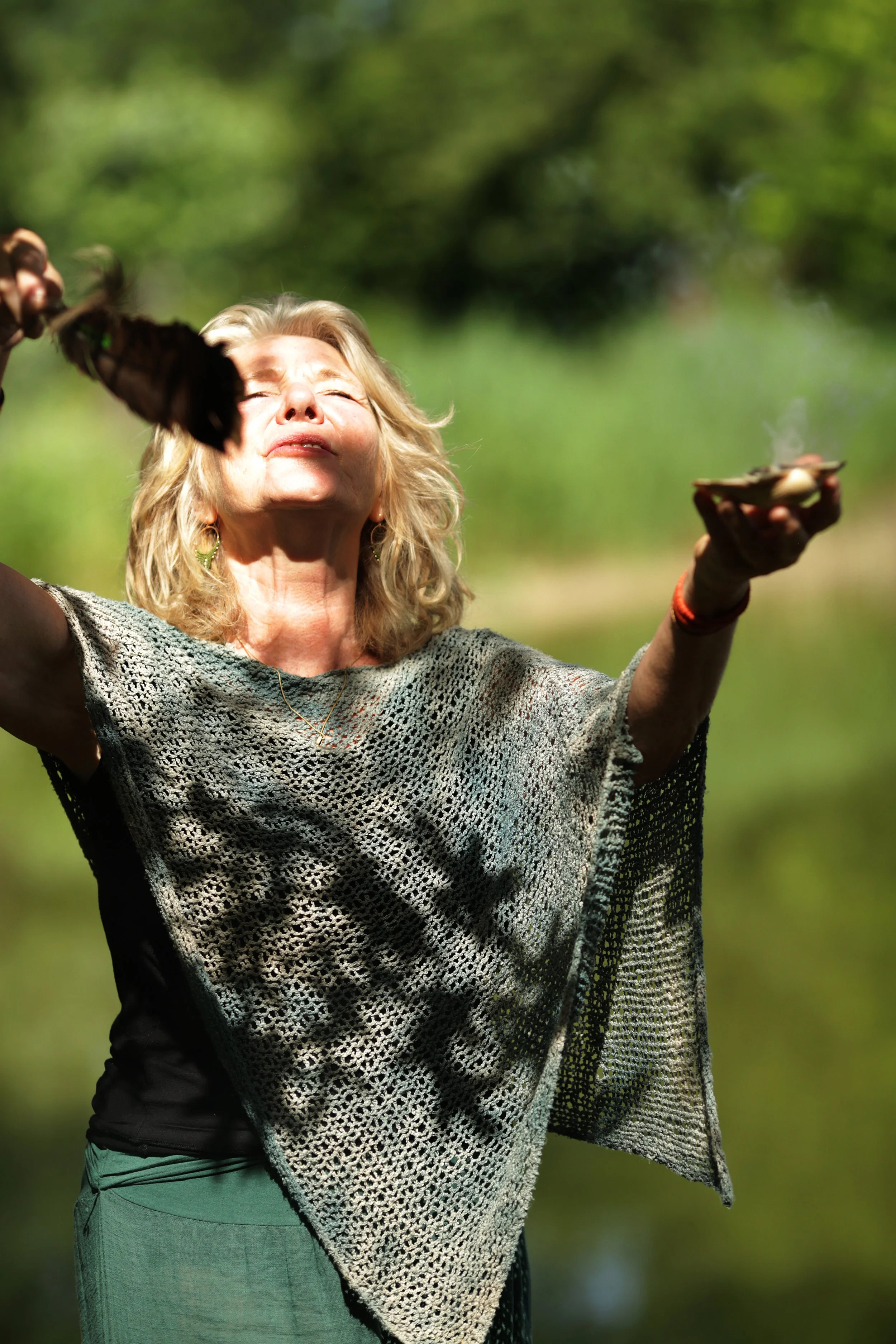 Een oudere vrouw met blond krullend haar staat buiten in de natuur, haar ogen dicht en haar gezicht omhoog gericht, terwijl ze haar armen in de lucht houdt en haar hand op haar hoofd legt, in de zon.