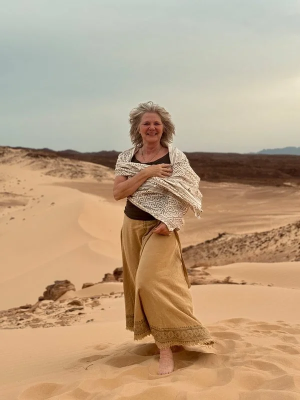 Vrouw glimlachend in de woestijn, gekleed in losse, beige kleding, wind in haar haar.