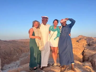 Vier mensen staan samen op een rotsachtige plek in een woestijnachtig landschap onder een blauwe lucht, lachend en poseren voor de foto.