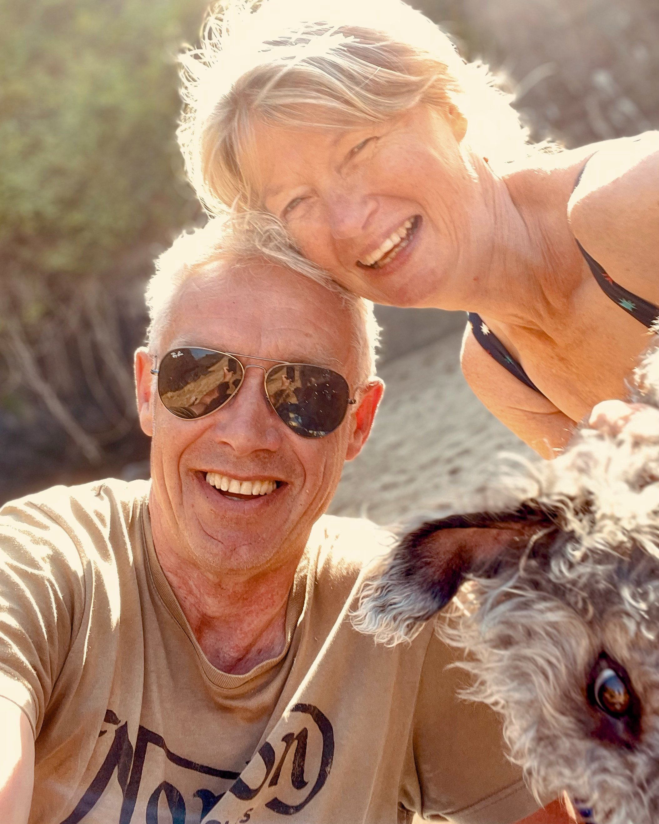 A smiling elderly couple taking a selfie outdoors with their dog, on a sunny day near a rocky or sandy area with trees in the background.