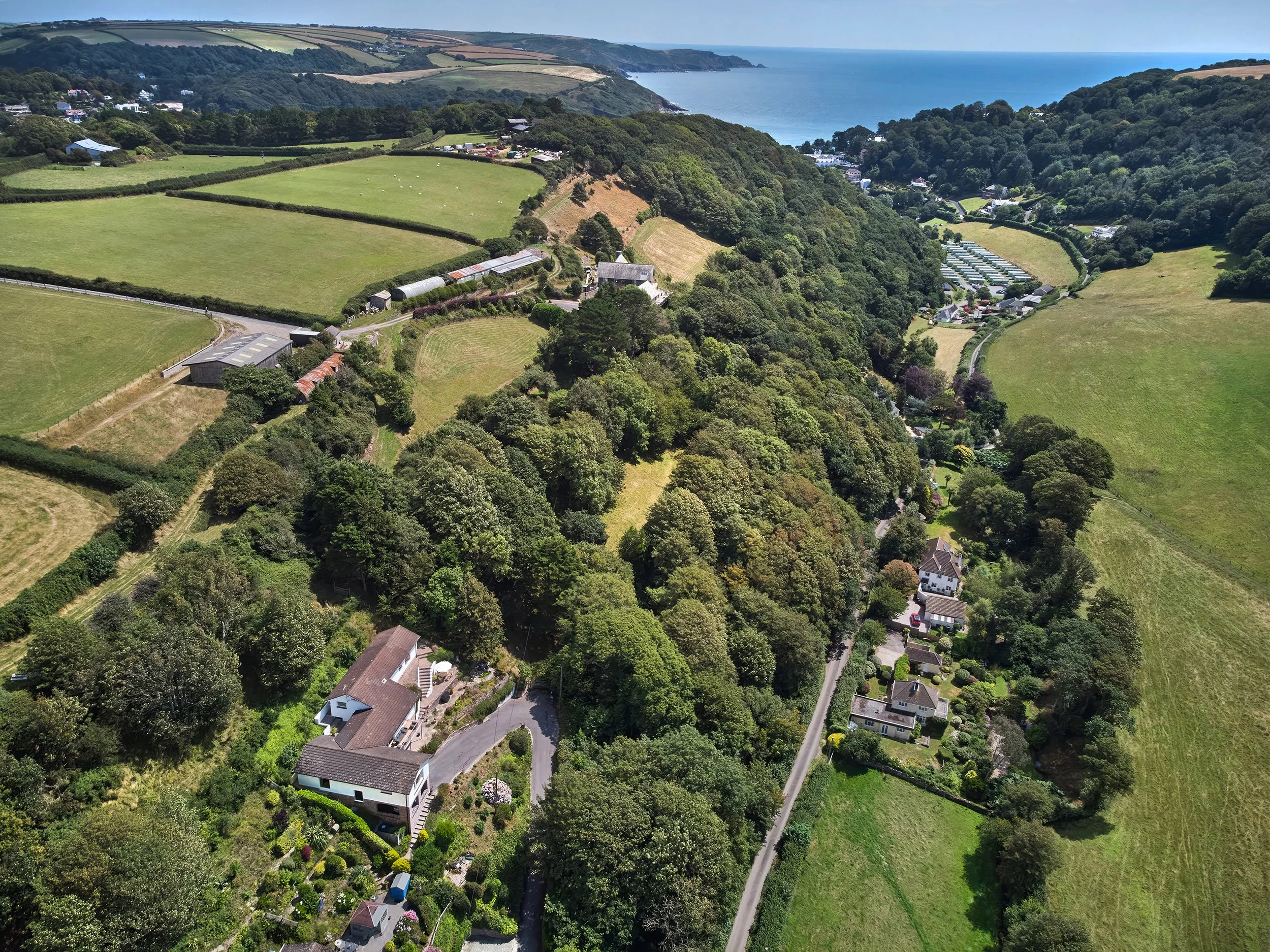 Aerial view of a rural landscape with houses, roads, wooded areas, and fields, featuring a coastline in the background.