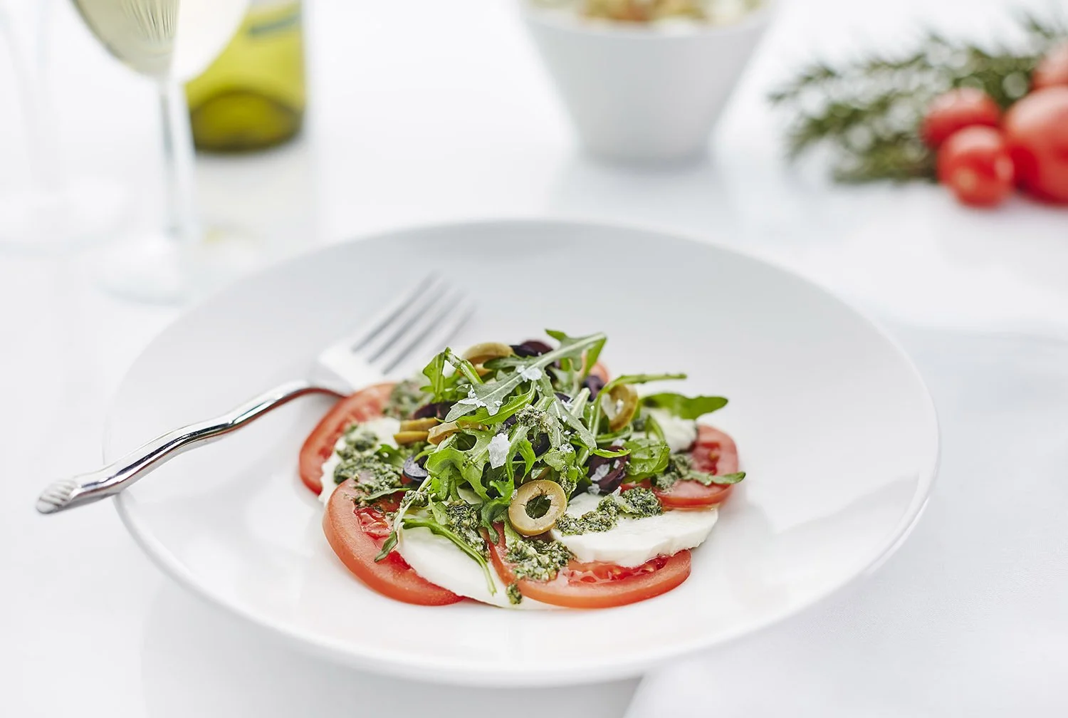Close-up of a fresh vegetable salad with tomatoes, olives, arugula, and cheese on a white plate, with a fork on the side.