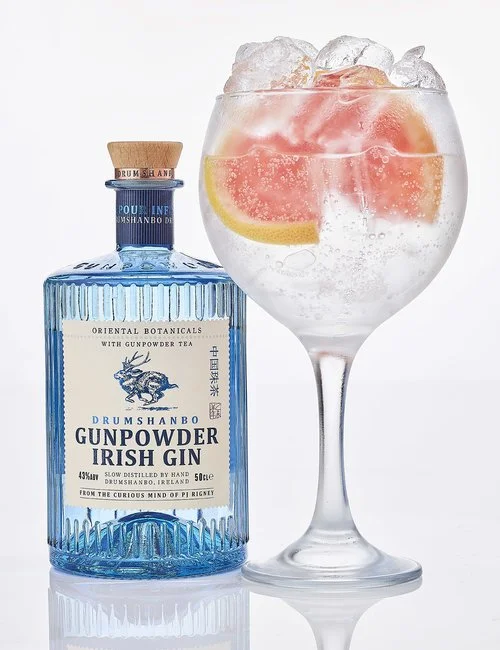 A bottle of Drumshanbo Gunpowder Irish Gin next to a glass of gin and tonic with ice and grapefruit slices.