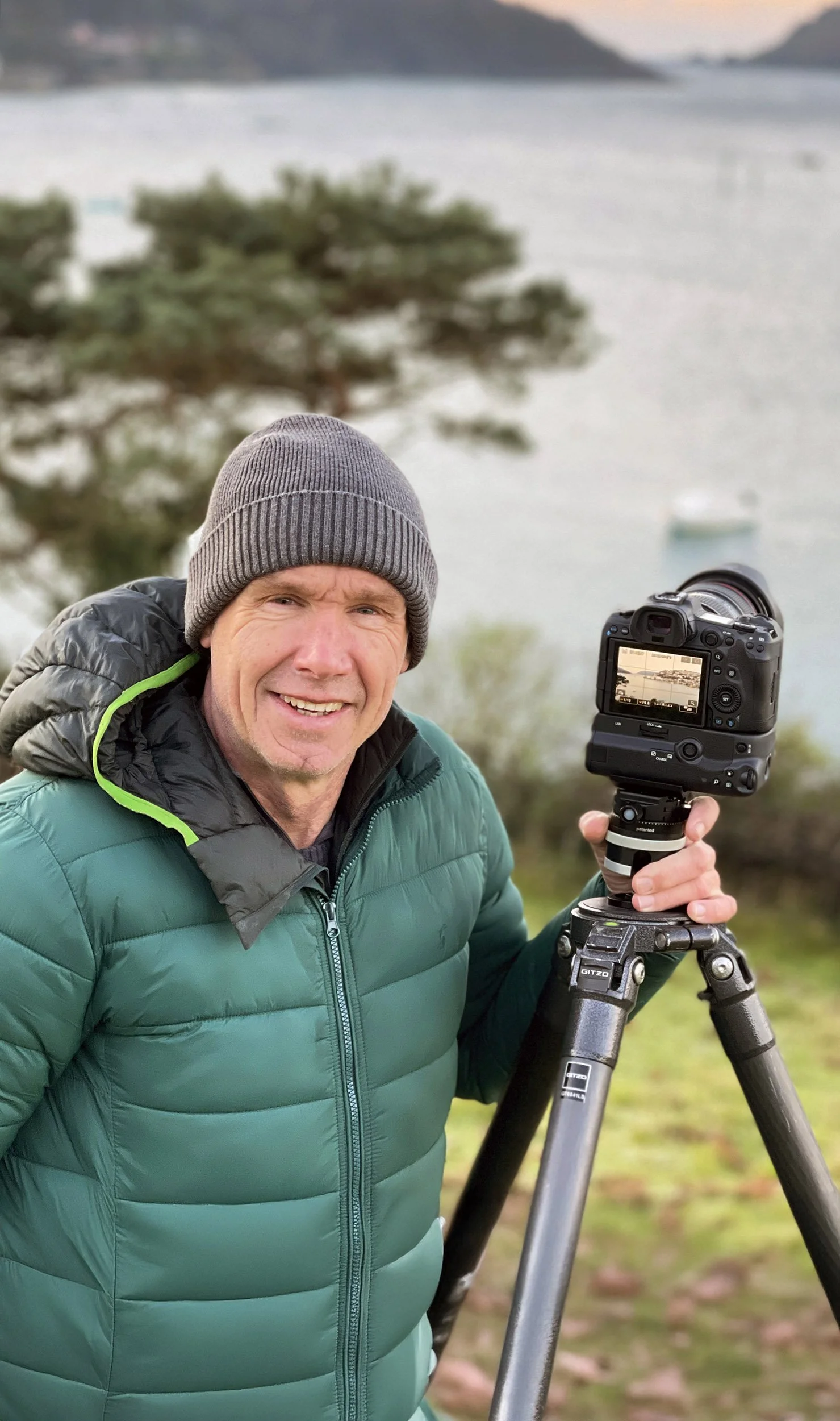 A man wearing a gray beanie and green padded jacket holding a camera on a tripod outdoors near a body of water with a boat, trees, and hills in the background.
