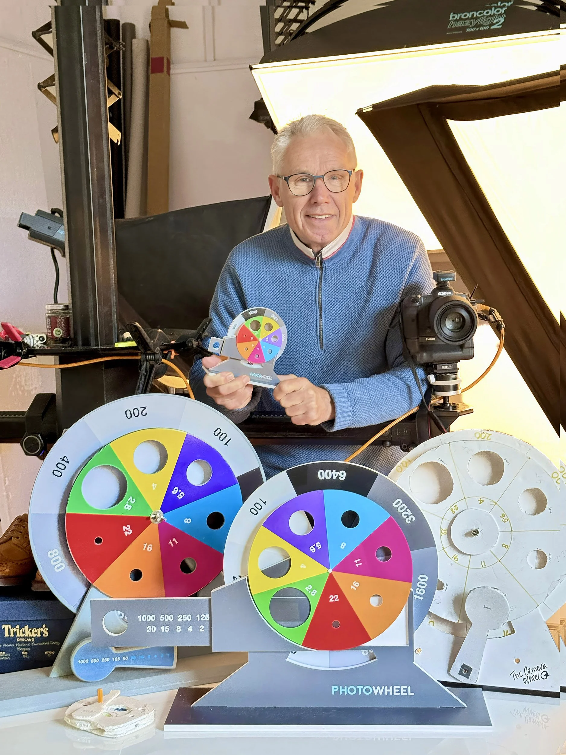 A man with glasses and gray hair in a blue sweater inside a photography studio, holding a colorful spinning wheel. Around him are several large color wheels with various colors and numbers, and a camera mounted on a stand to his right. Studio lighting equipment is visible above.