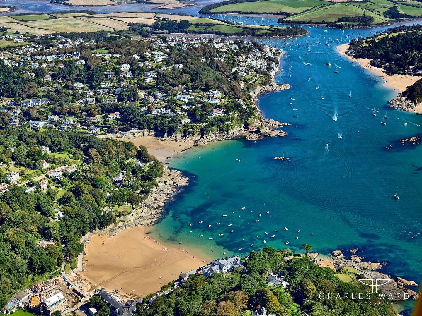 Aerial view of Salcombe,  a coastal town with beaches, houses, and numerous boats on the water, surrounded by green hills and farmland.
