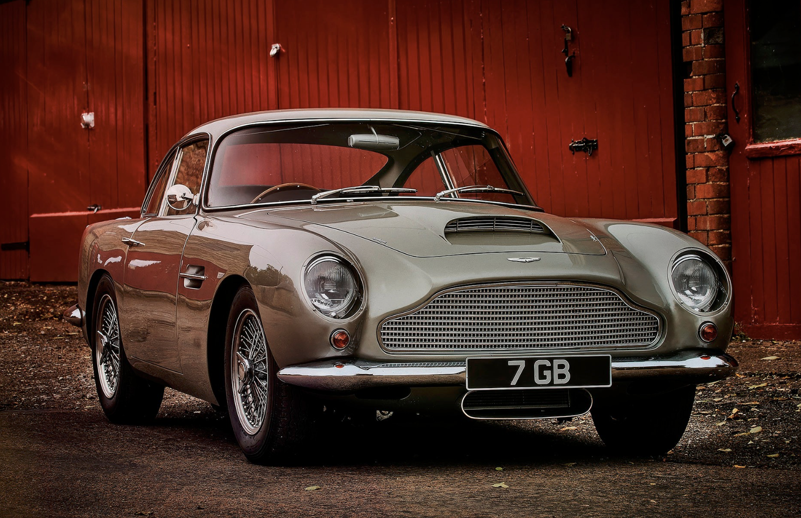 A classic silver Aston Martin DB4 GT sports car parked in front of a red wooden garage door.