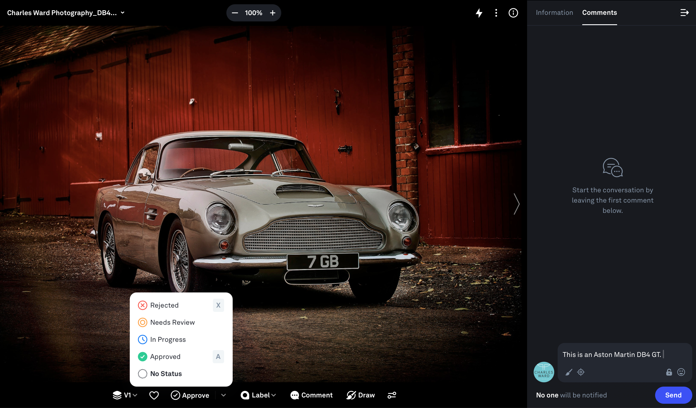 A classic silver Aston Martin DB4 GT parked in front of a red wooden building with a brick column.