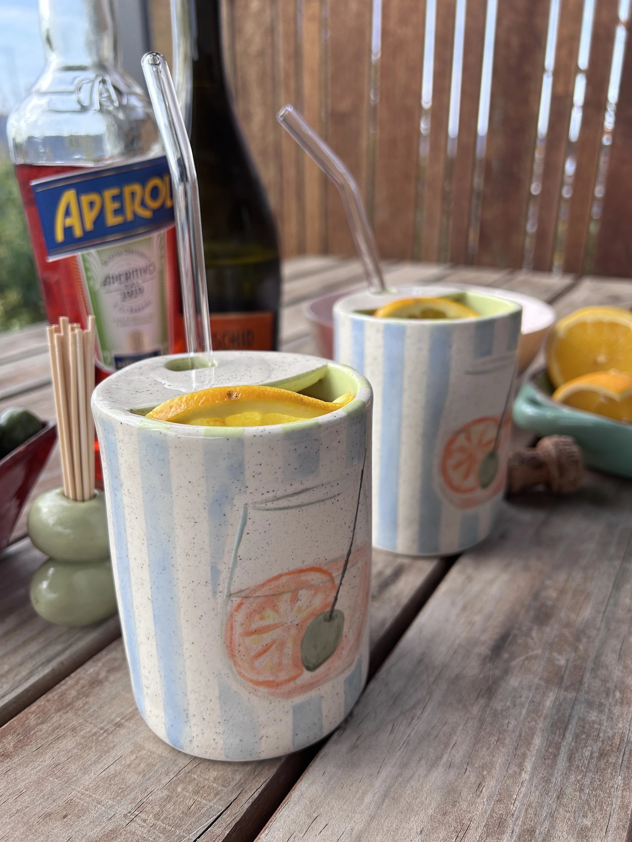 Two paper cups with slices of lemon inside, placed on a wooden table outdoors. Each cup has a straw, and there are other beverages and condiments in the background.