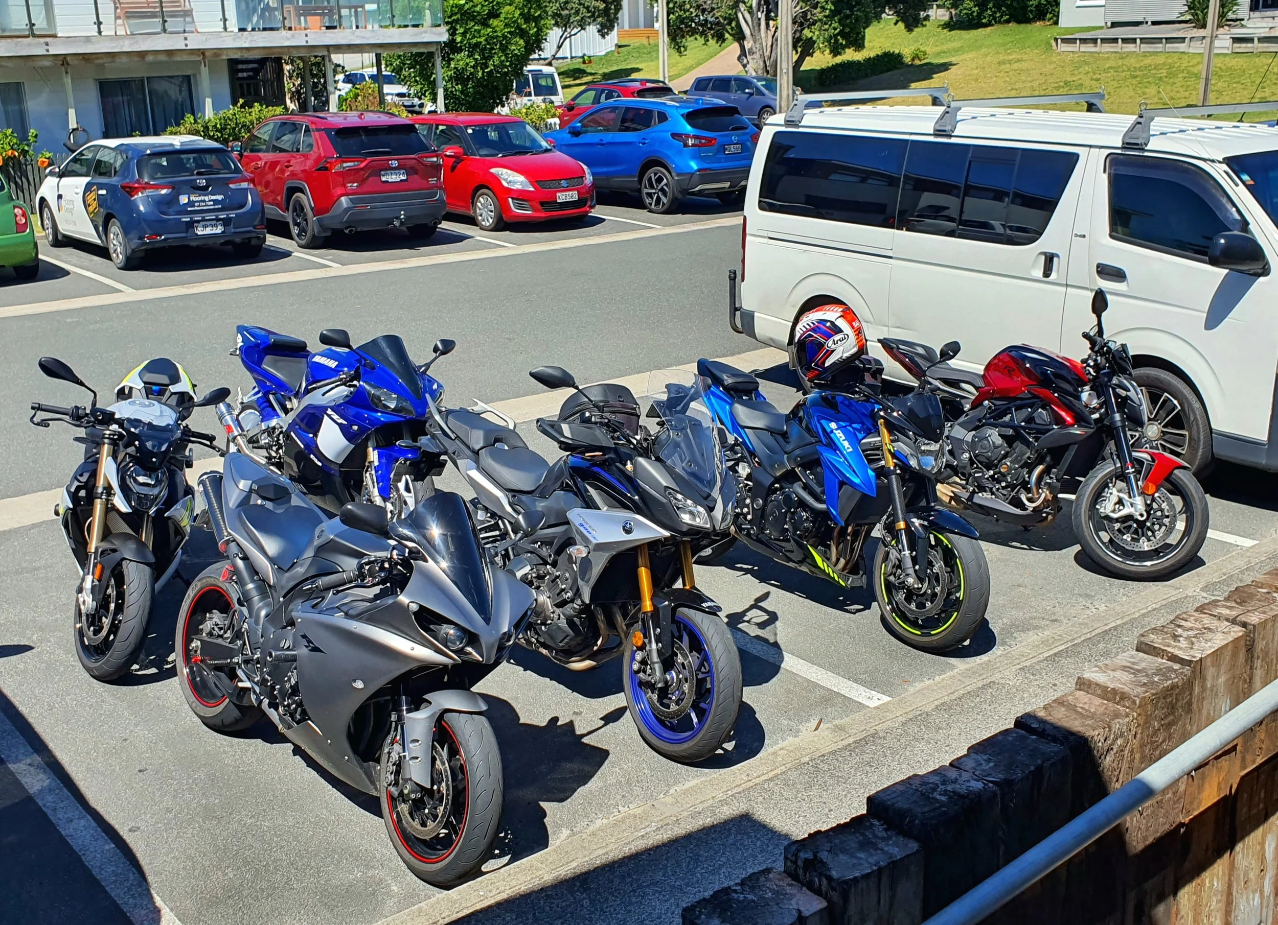 Multiple motorcycles parked in a parking lot in the foreground, with several cars parked behind them including a blue, a red, and a white vehicle. A white van with roof rack and a motorcycle helmet on it is visible. In the background, there are buildings, green trees, and a pathway.