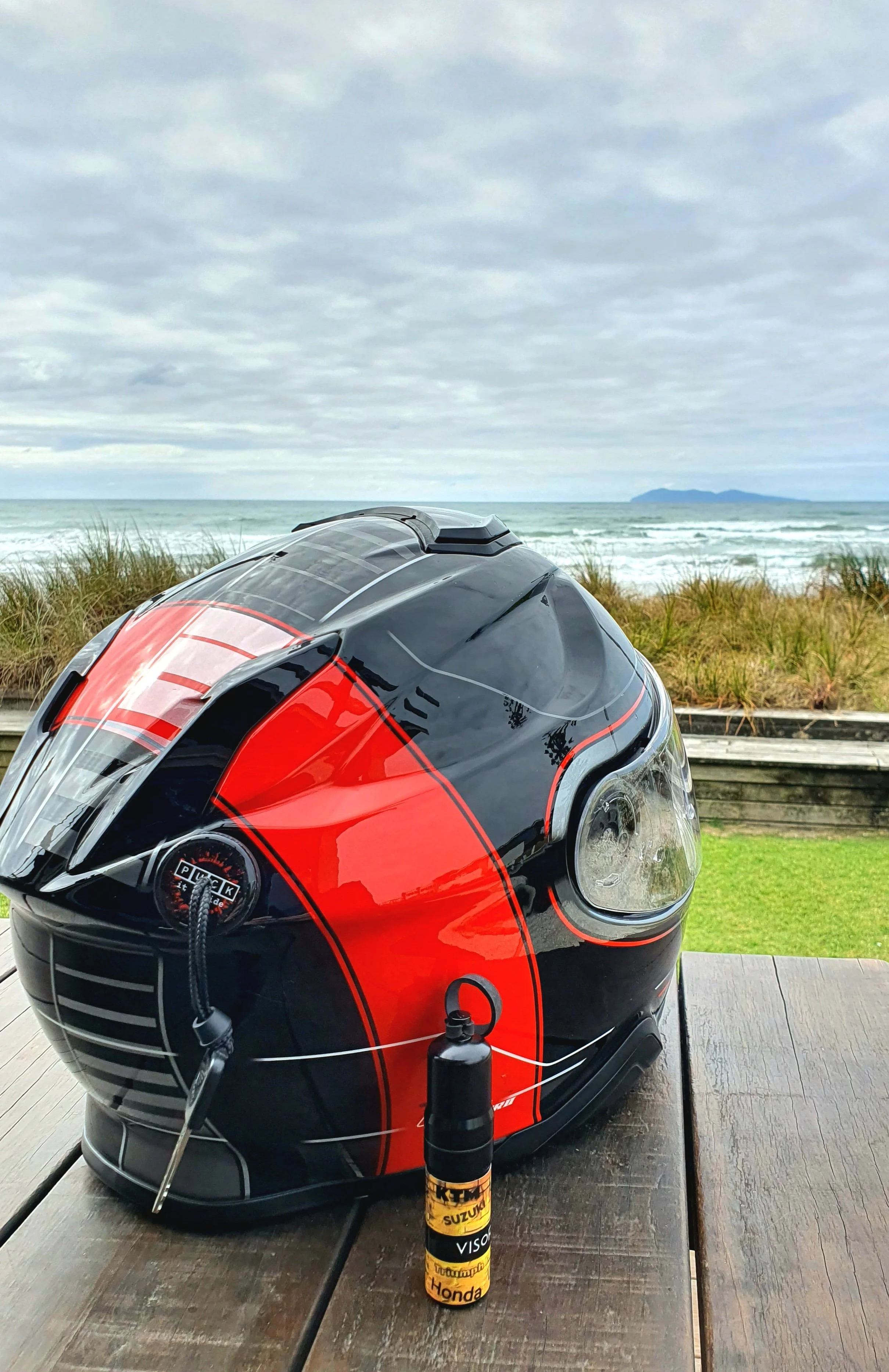 A motorcycle helmet with black, red, and gray design on a wooden outdoor table, with a yellow bottle labeled KTM, Suzuki, Honda, and Visor standing upright in front of it, a cloudy sky, and the ocean with a distant landmass in the background.