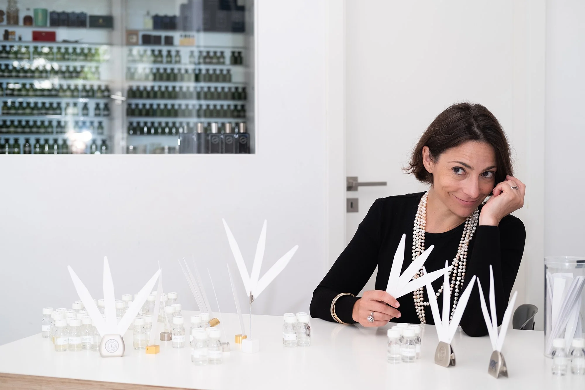 A woman with dark hair wearing a black top and pearl necklace, smiling and holding white paper sticks, sitting at a table with bottles of perfume and white display stands.