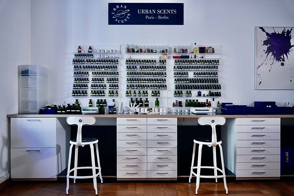 A display of essential oils or fragrances on white shelves, a sign reading 'Urban Scents Paris - Berlin,' and a workspace with chairs, drawers, and containers in a clean, modern room.