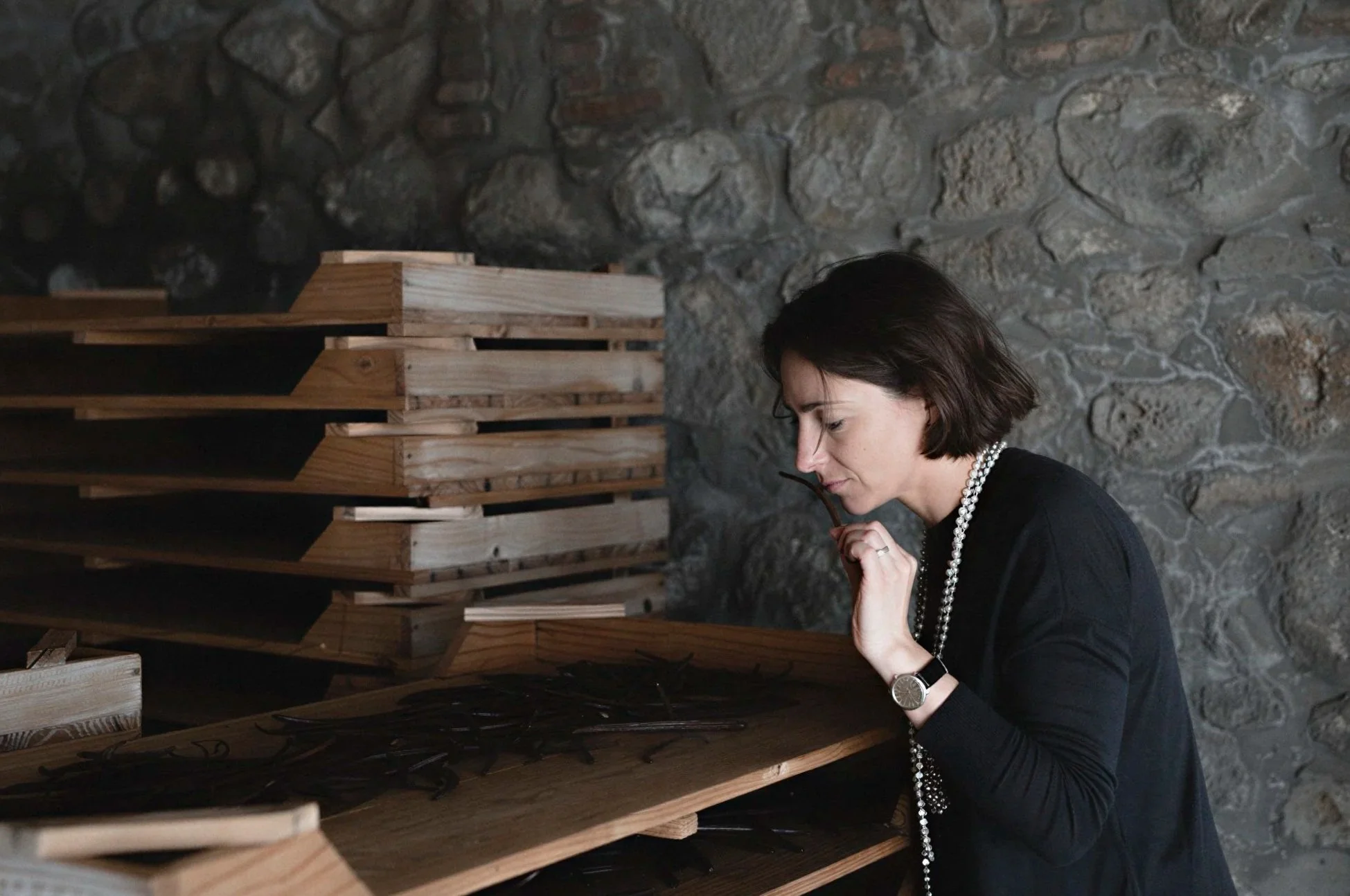 A woman with short dark hair, wearing a black top, pearl necklace, and a watch, is standing in front of a stone wall and looking at a wooden structure with black wire or cables hanging from it.