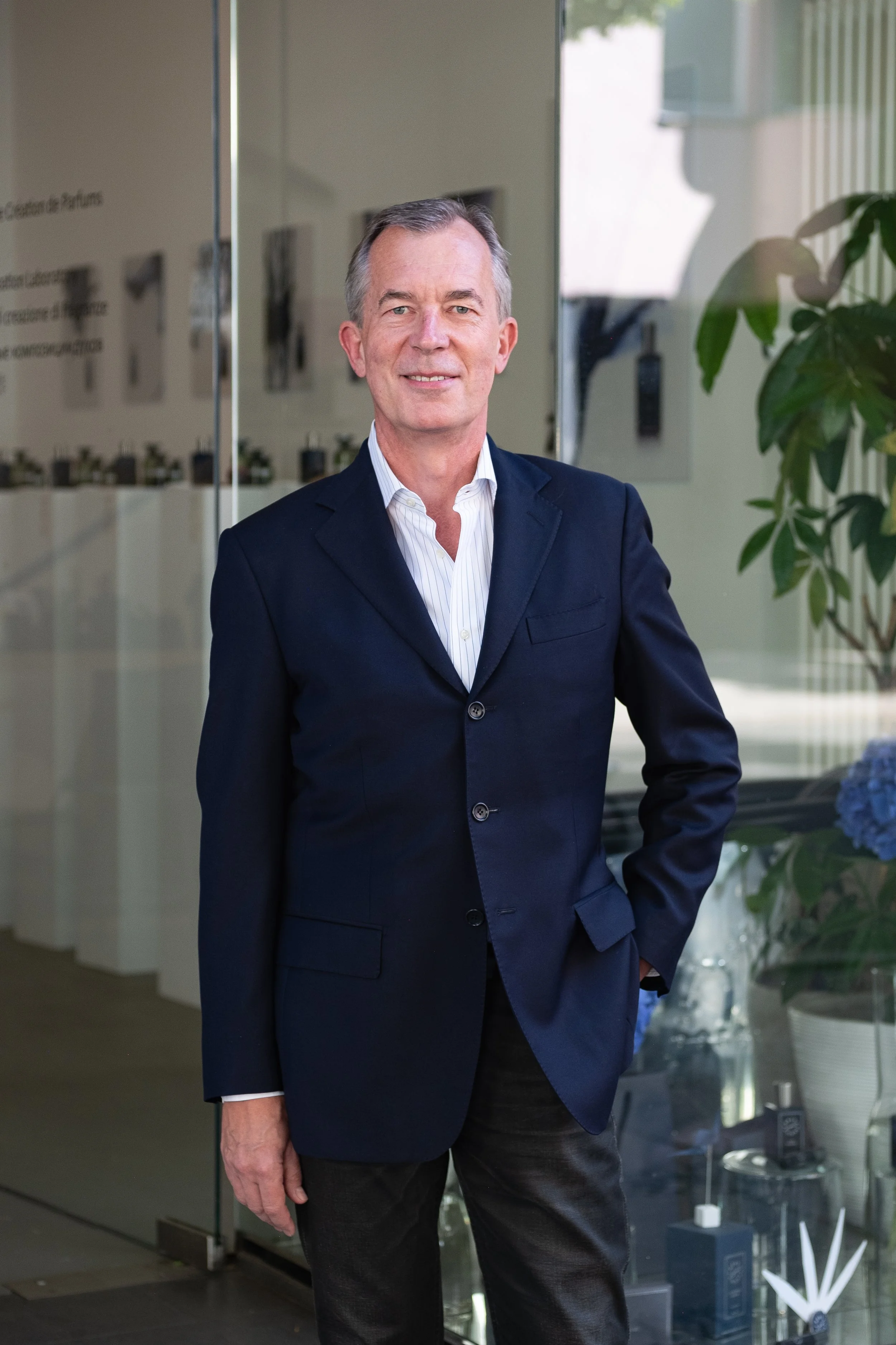 A middle-aged man dressed in a dark navy suit and white shirt standing inside an office building with glass walls. Behind him, there are potted plants and some framed pictures or documents on the wall.