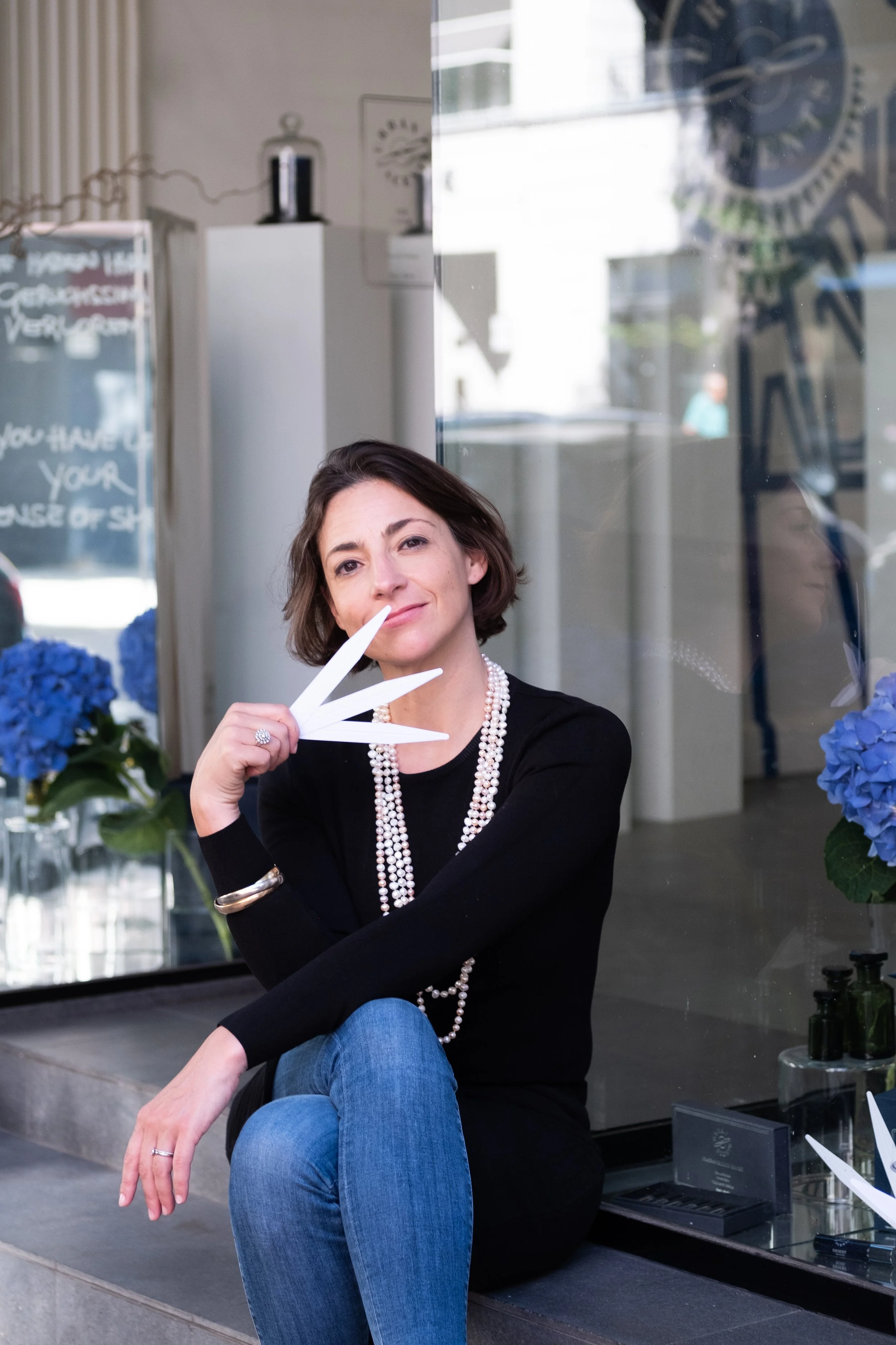 A woman with short brown hair and pearl necklace sitting outside a storefront, holding a white paper fan near her face. She is wearing a black top, blue jeans, and has rings and a bracelet. Blue hydrangeas are visible inside the store.