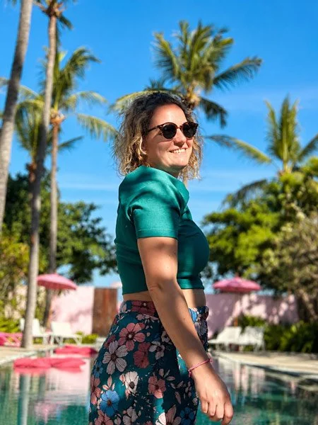 Cyrielle Patys, femme souriante portant des lunettes de soleil, un haut vert et une jupe à fleurs, debout près d'une piscine avec des palmiers en arrière-plan, par une journée ensoleillée.
