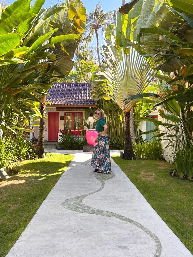 Cyrielle Patys, femme portant une jupe florale et un haut vert pose sur un chemin sinueux dans un jardin tropical avec des palmiers