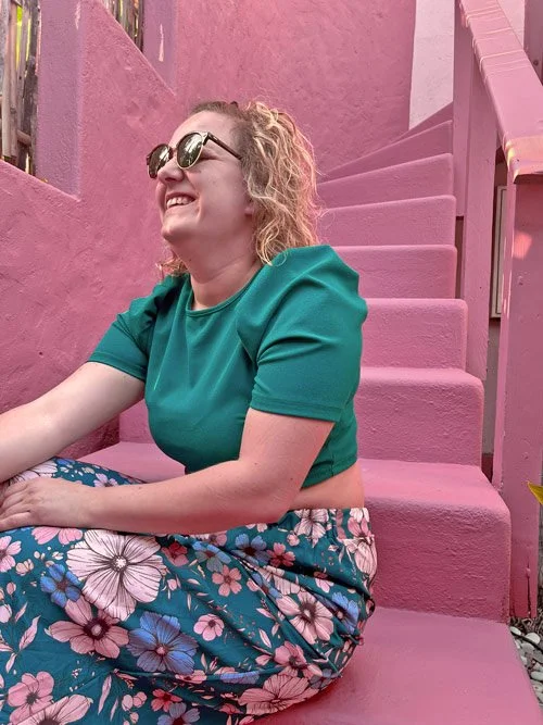 Cyrielle Patys, femme souriante portant des lunettes-de-soleil, un haut vert et une jupe à fleurs, assise sur un escalier rose dans un décor coloré.