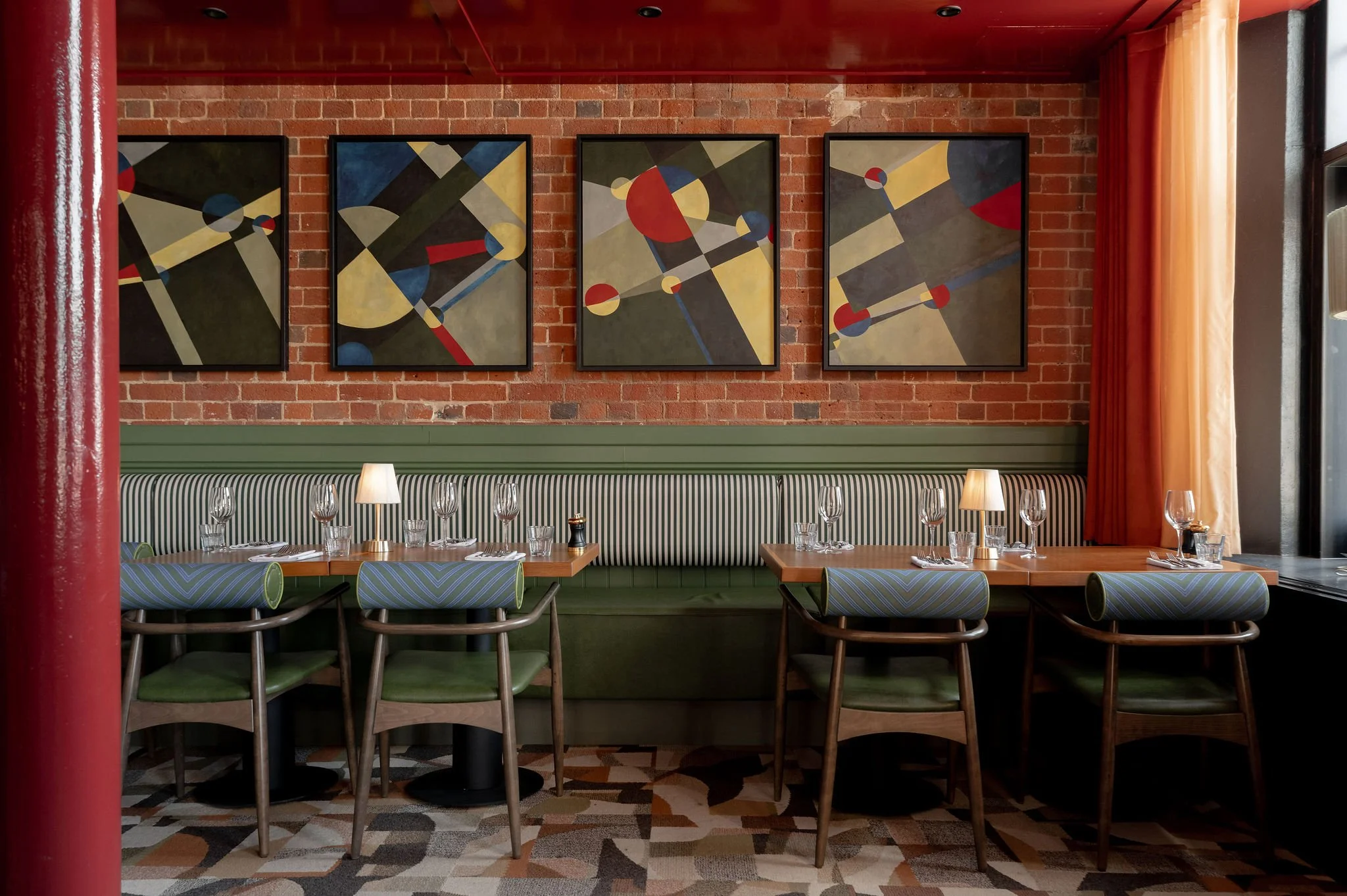 Interior of a restaurant with a red brick wall, colorful geometric abstract paintings, a long green-striped booth, tables with glasses and cutlery, and patterned carpet flooring.