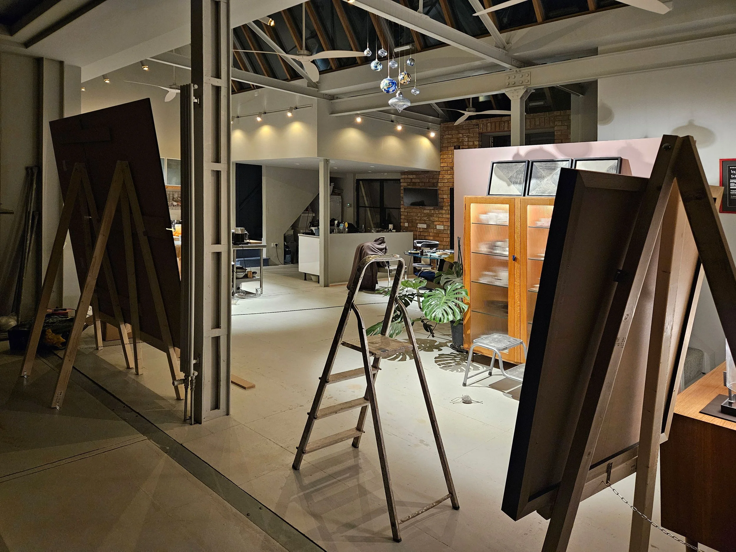 Interior of an art gallery or studio with multiple paintings and framed artwork displayed on easels and walls. The space features a high, vaulted ceiling with exposed beams and hanging spherical decorations. Construction tools and chairs are present, indicating ongoing setup or renovation.