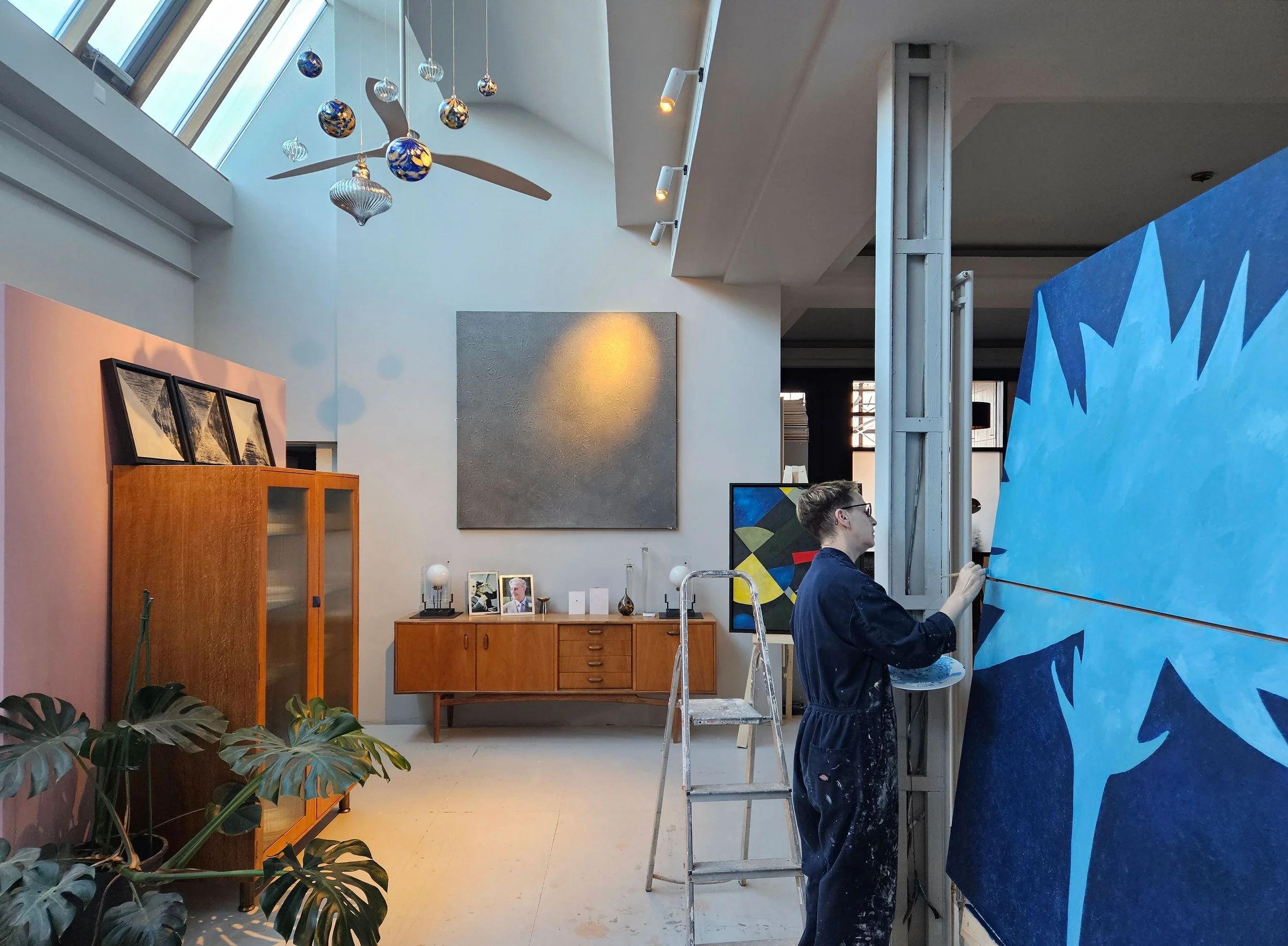 An art gallery interior with a person painting a large blue canvas with star shapes. The gallery has modern decor, artworks on the walls, and decorative ornaments hanging from the ceiling.
