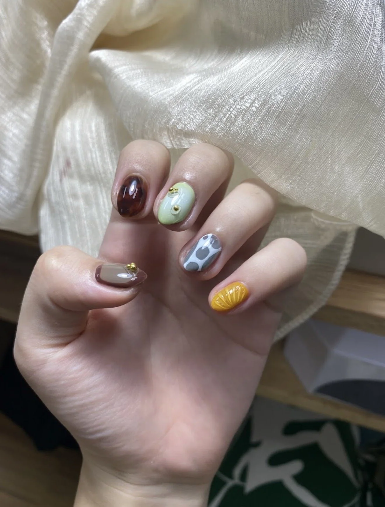 Hand with various artistic nail designs, including a brown and black swirl, pastel green with small gold accents, blue with white and gray spots, beige with a small gold bead, and yellow with a textured fan-like pattern, resting on cream-colored sati